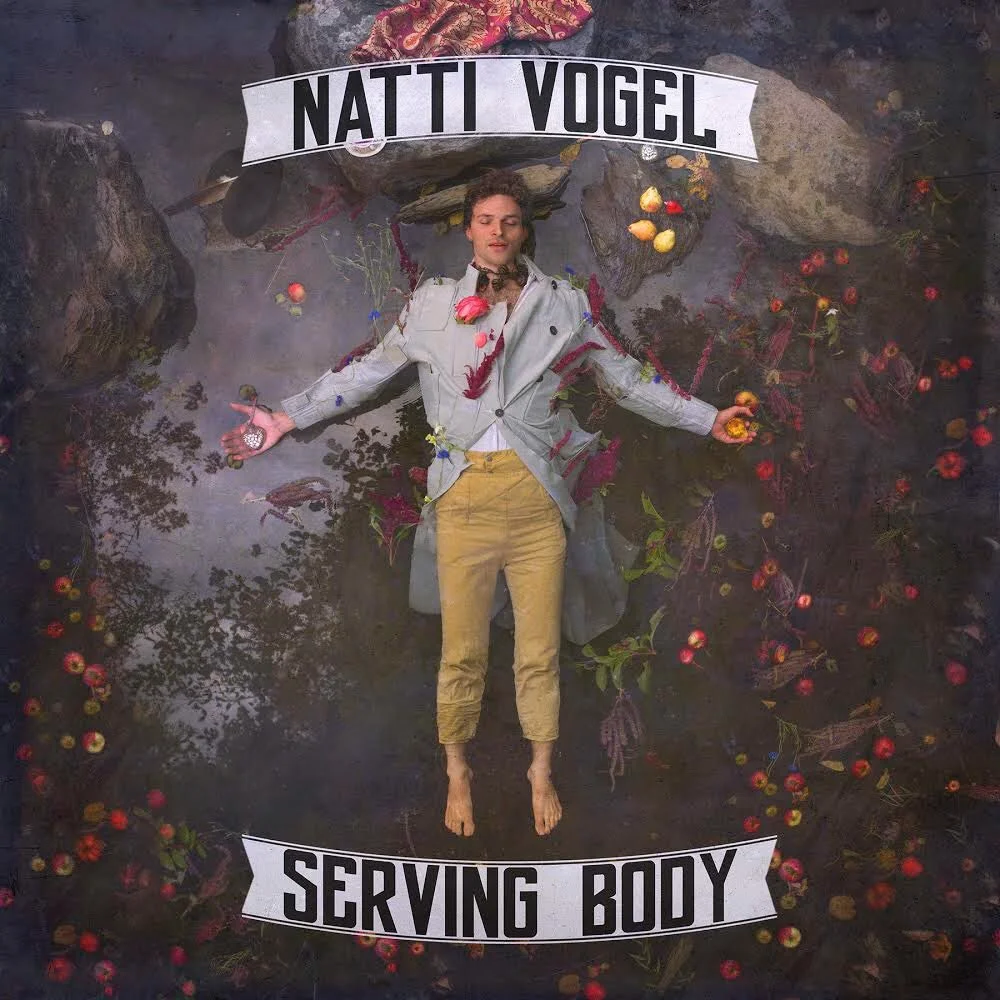 Natti Vogel lying on the ground with flowers, fruits, and leaves scattered around, dressed in a beige coat and yellow pants, with a sign above reading 'Natti Vogel' and another below reading 'Serving Body'.