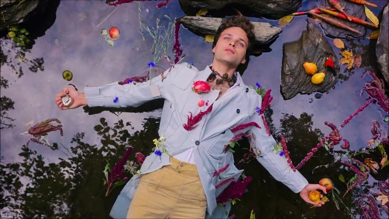 Natti Vogel with dark curly hair lying in a body of water surrounded by colorful flowers and rocks. He is wearing a light gray jacket and beige pants, holding an apple in each hand, looking up with a contemplative expression.