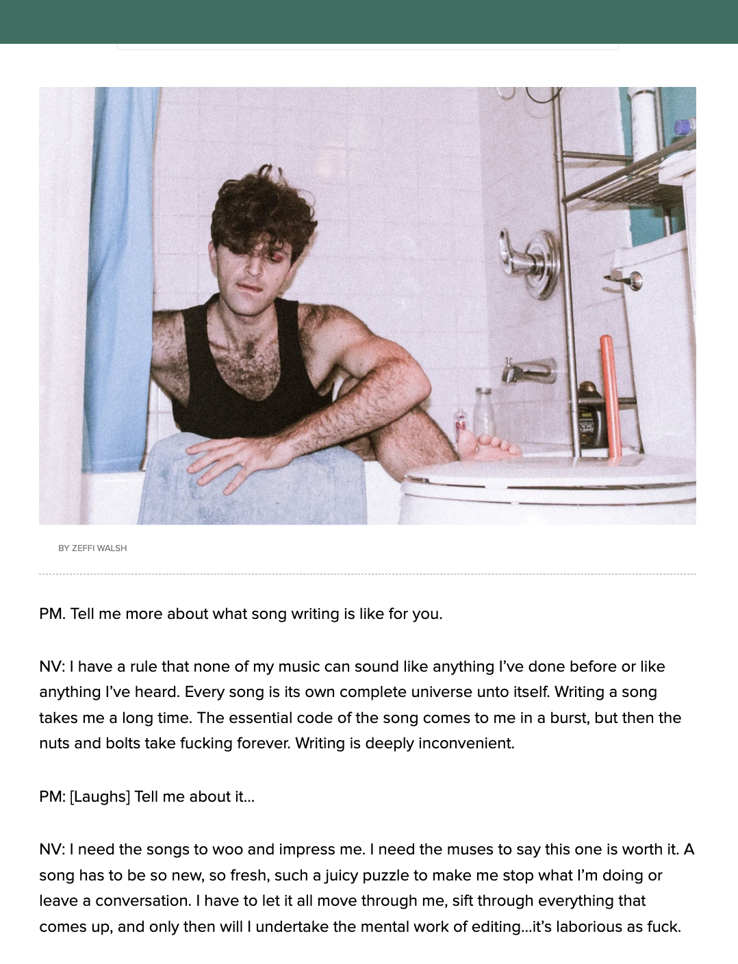A young man with dark, curly hair and a black tank top sitting on the edge of a bathtub in a bathroom. He appears to be in thought, with a somewhat serious expression. The bathroom has white tiled walls, a shower head, a bottle of soap, a red object,