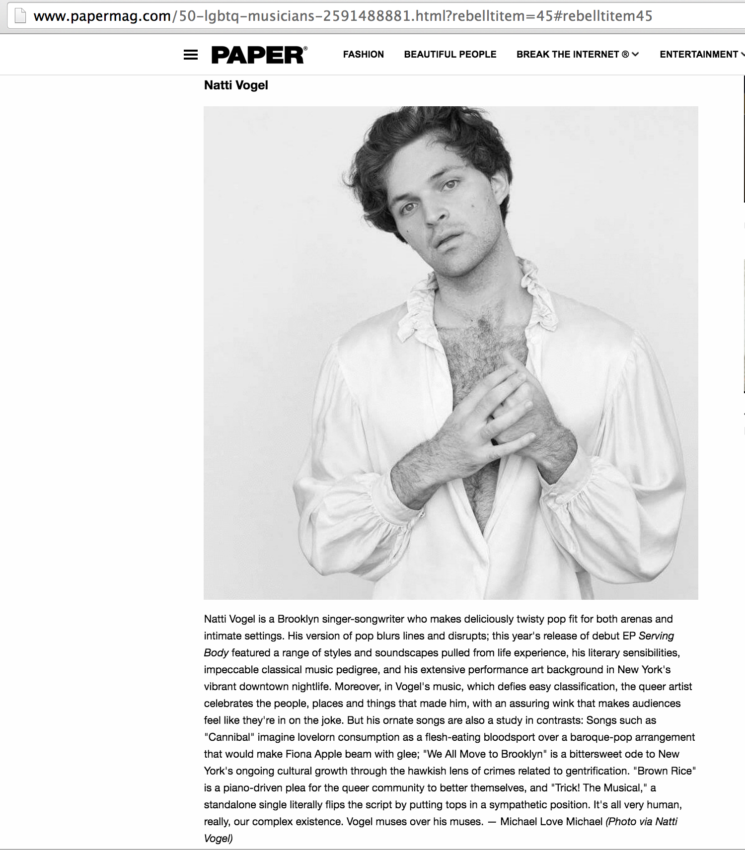 Black and white portrait of Natti Vogel, with wavy hair, wearing a ruffled shirt, and touching his chest with one hand. From PAPER Magazine