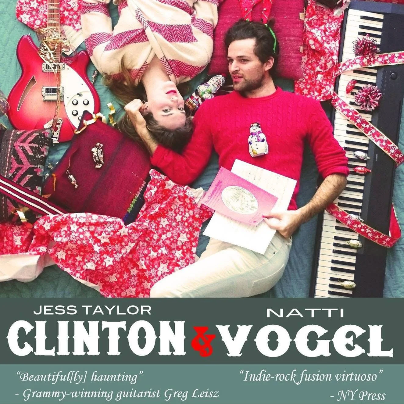 A man and woman lying on the floor surrounded by Christmas-themed gifts and decorations. The woman is looking up with her head on a pillow, and the man is holding a pink greeting card. There is a red guitar, a toy snowman, a keyboard, and various wra