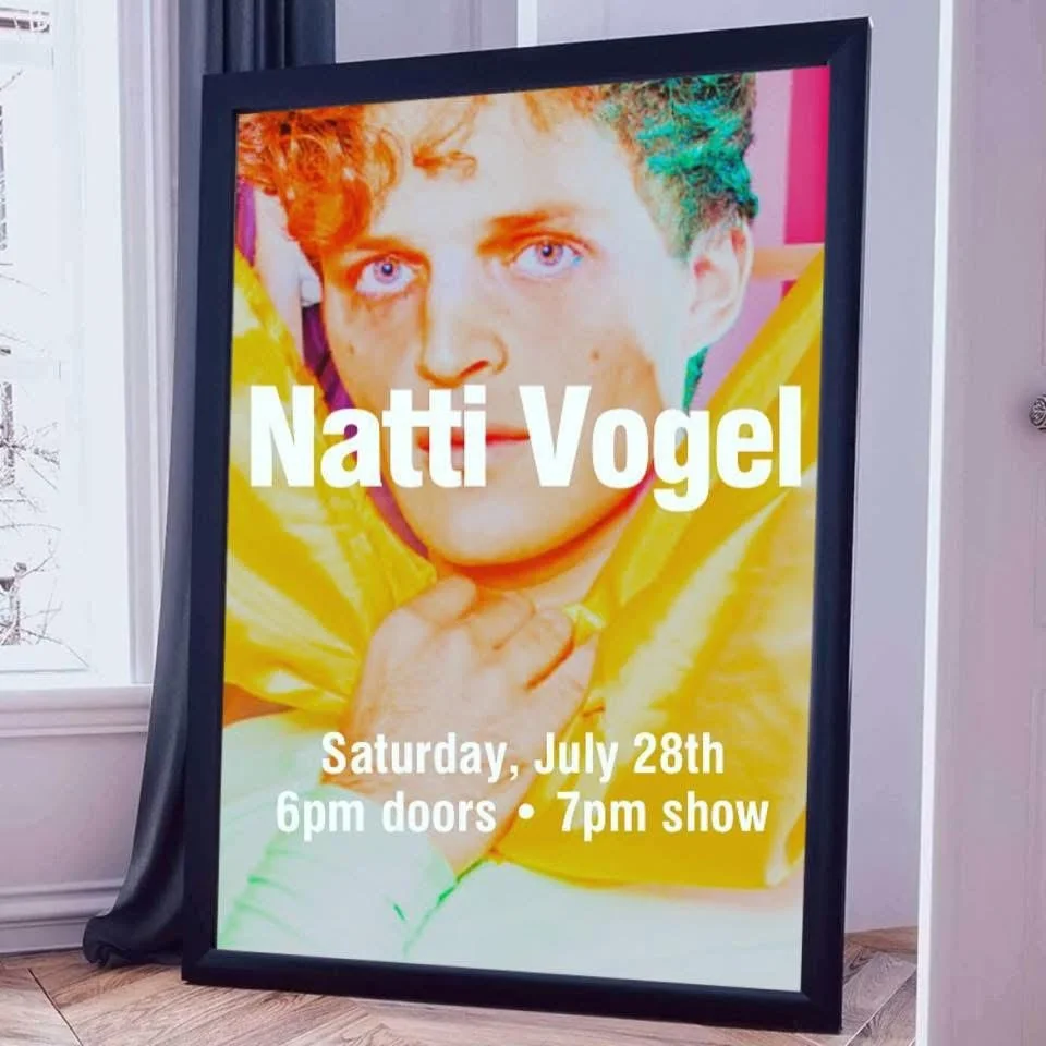 A framed poster promoting a performance by Natti Vogel, scheduled for Saturday, July 28th, with doors opening at 6 pm and the show starting at 7 pm. The poster features a bright, colorful portrait of Natti Vogel with blue eyes, curly hair, and a yell