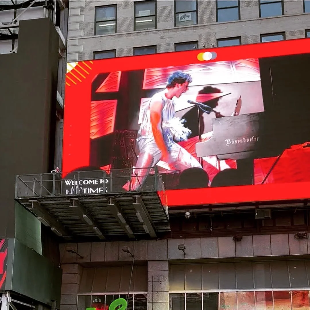 Large digital billboard in Times Square showing Natti Vogel playing a grand piano on stage, with a red background and stage lighting.