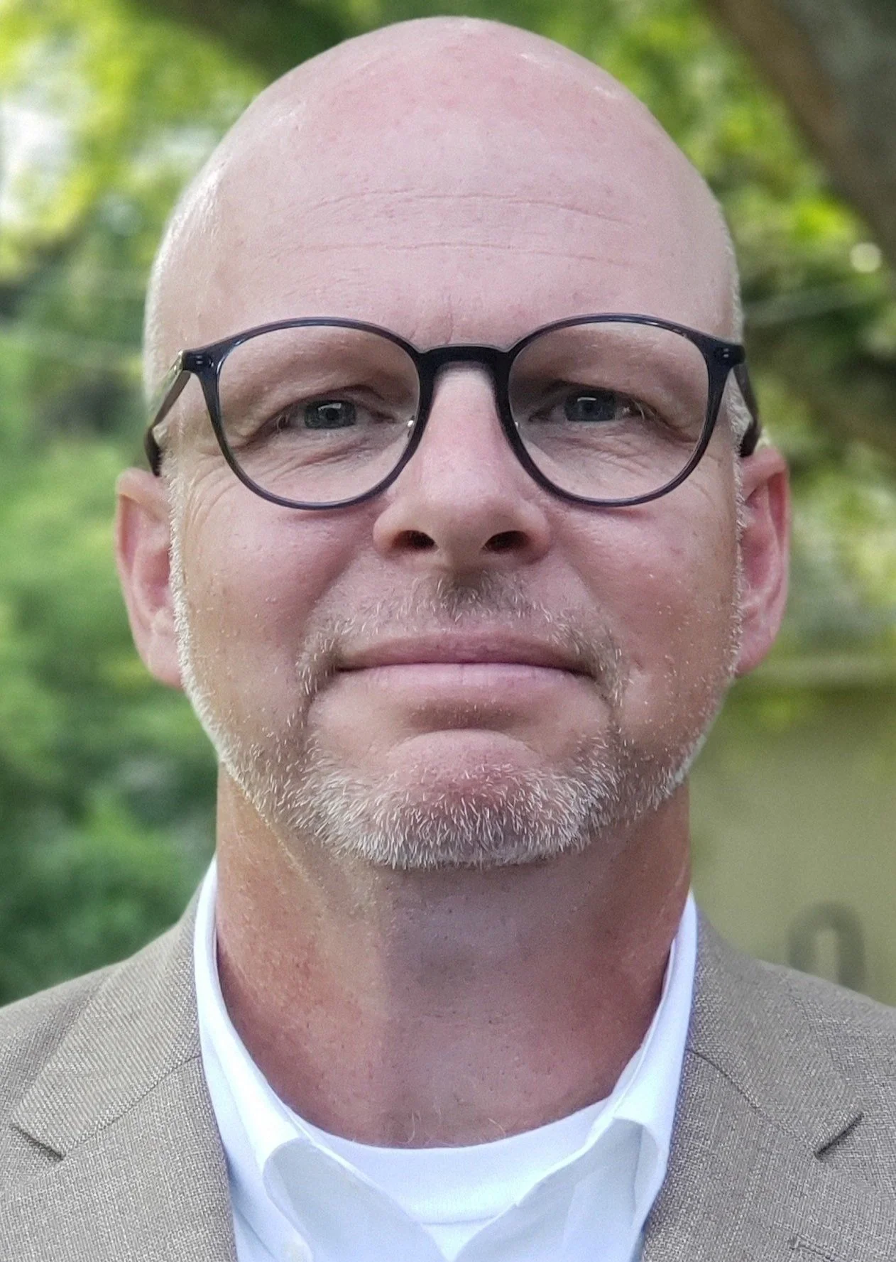 Close-up of a bald man with glasses, wearing a beige blazer and white shirt, outdoors with blurred green foliage in background.