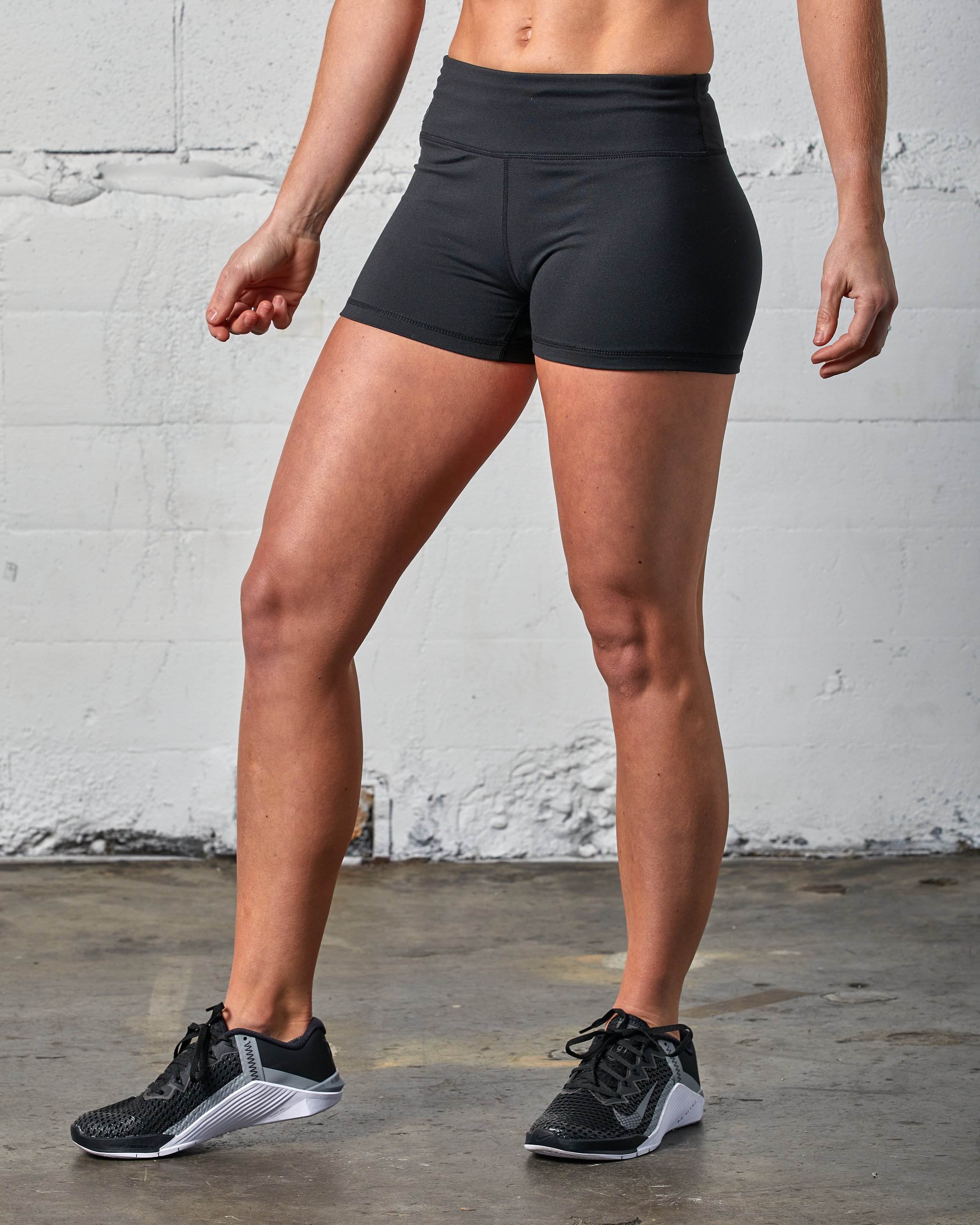 A woman wearing black athletic shorts and black athletic shoes, standing against a white wall with a concrete floor.