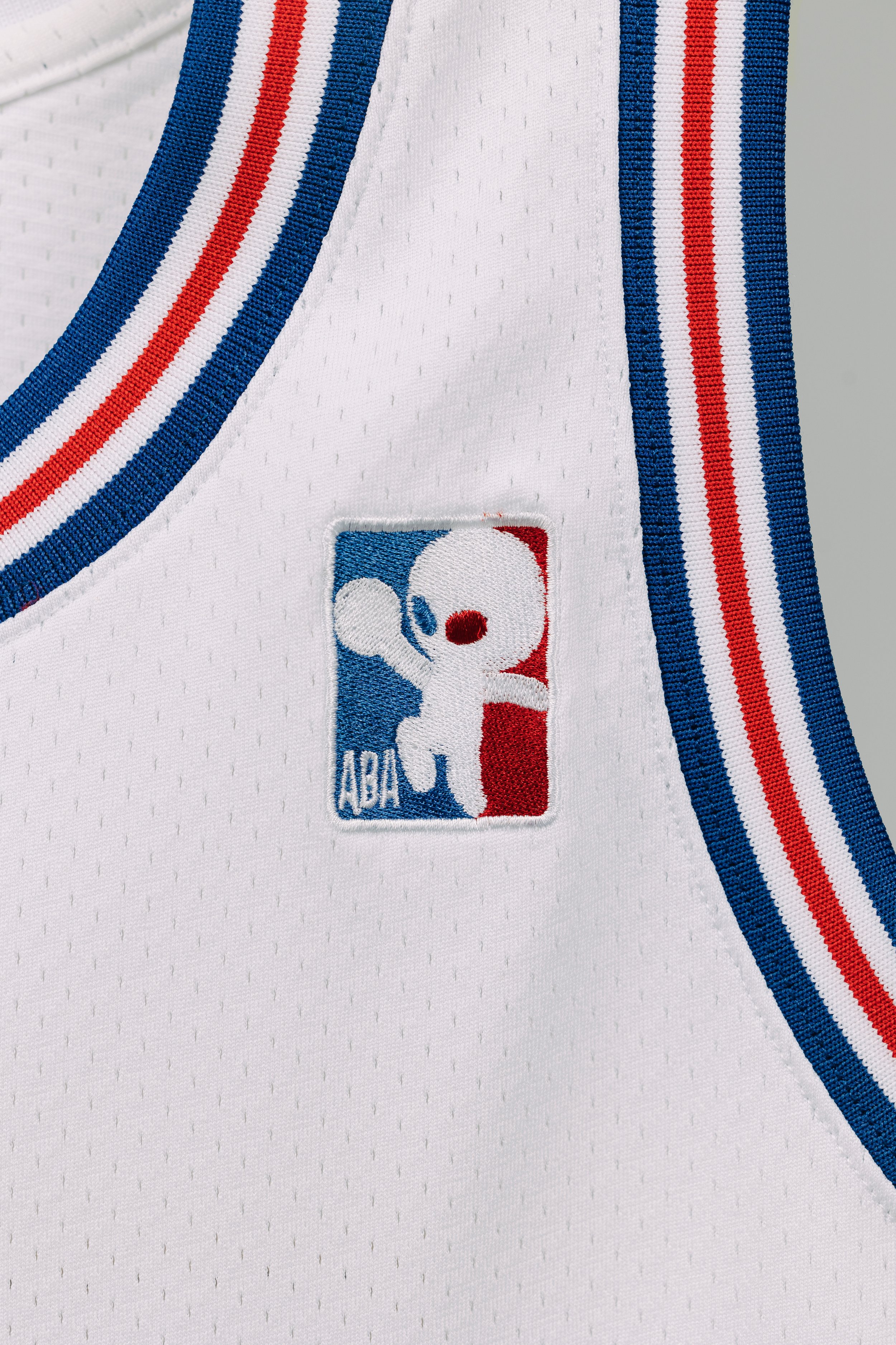 Close-up of a white NBA basketball jersey with the NBA logo patch showing a cartoon dog in red, white, and blue colors, with a blue and red striped trim.
