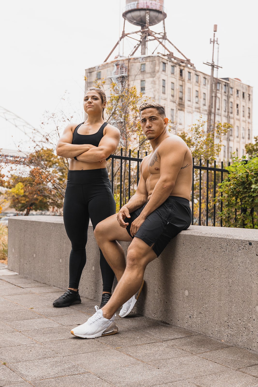 A muscular woman and a shirtless man with tattoos pose outdoors in athletic clothing, with a cityscape featuring a water tower and old buildings in the background.