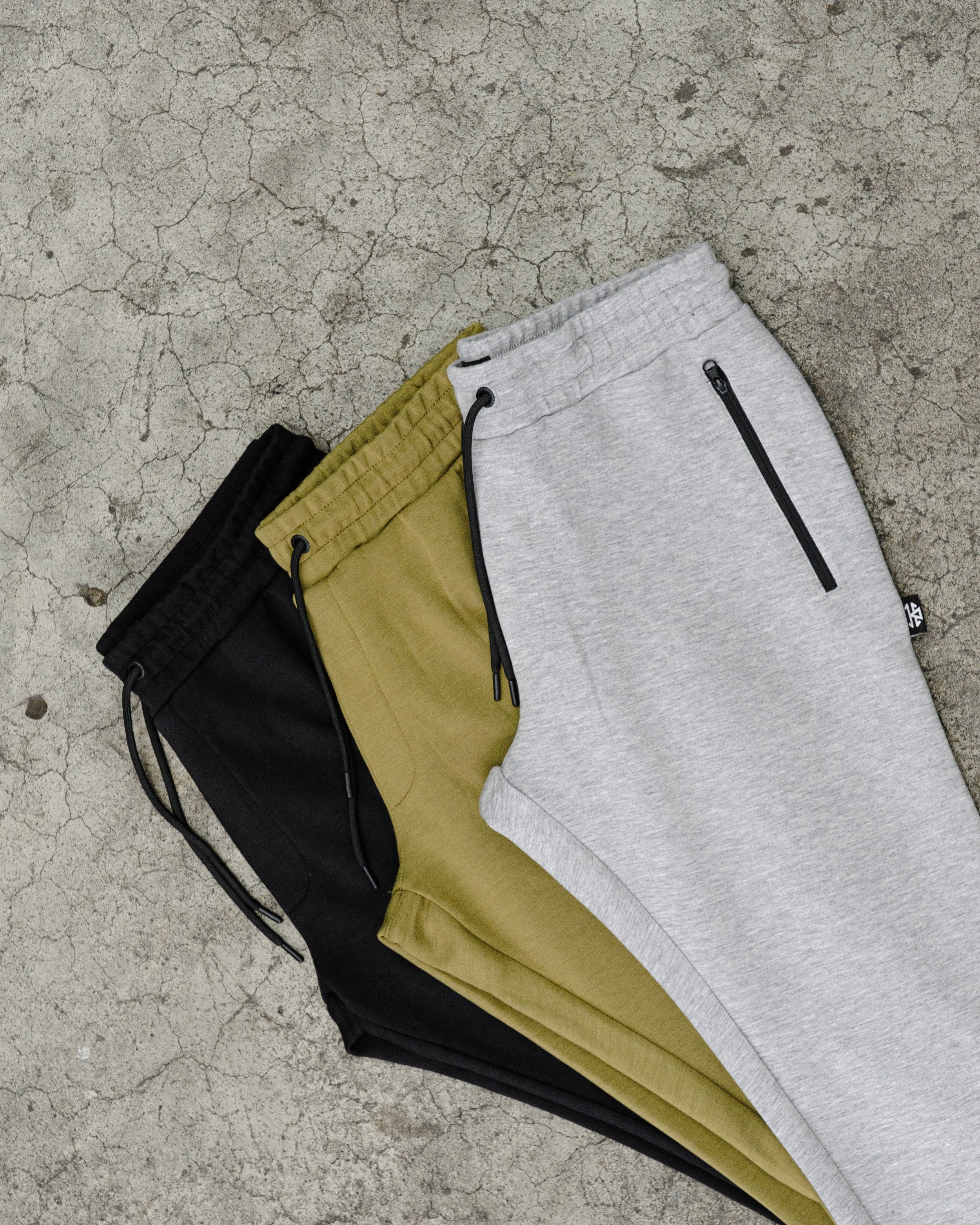 Three pairs of sweatpants in black, olive green, and light gray with zippered pockets and drawstrings, laid flat on a cracked concrete surface.