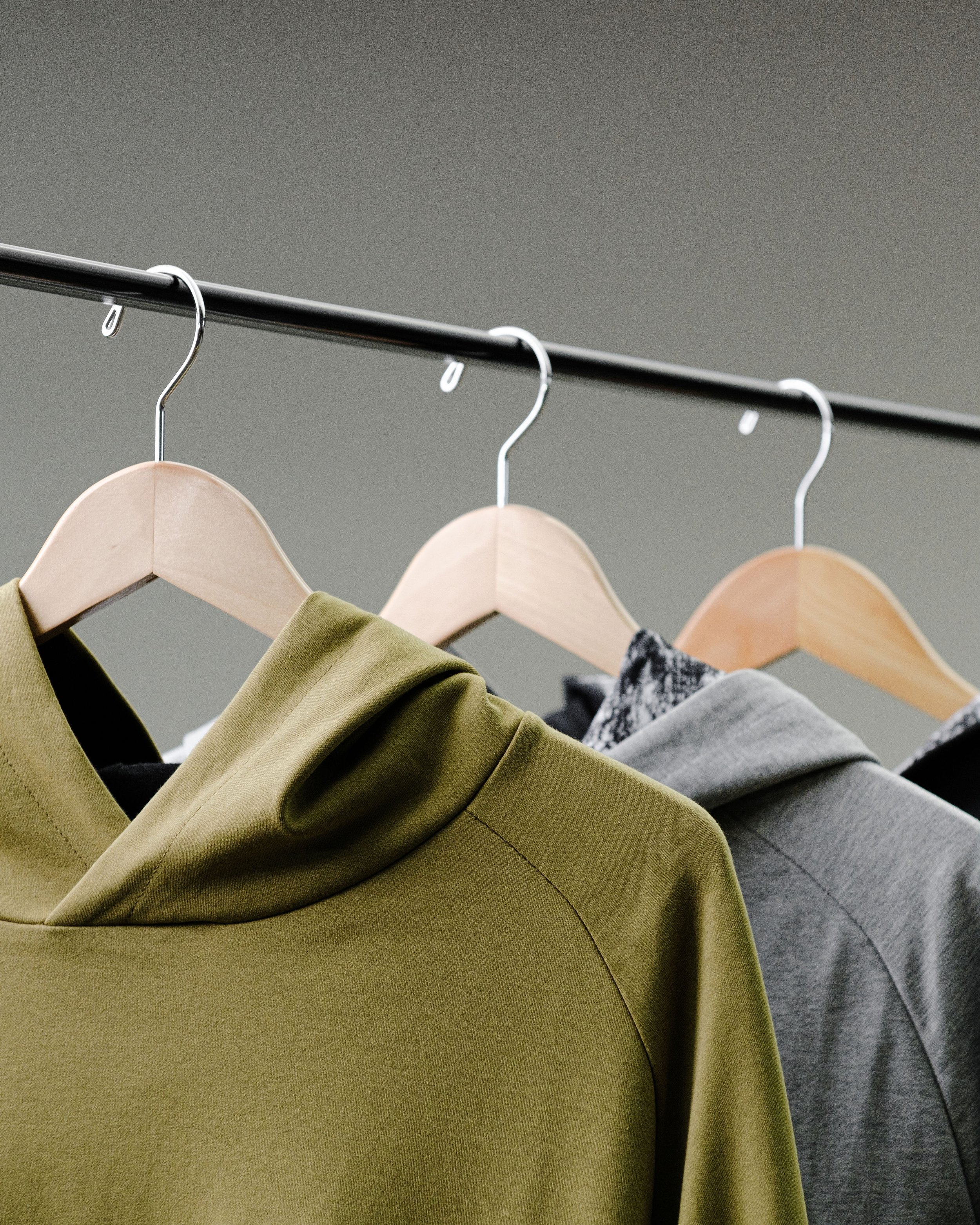 Three hoodies hanging on wooden hangers on a metal clothing rack, in olive green, black with grey marbled pattern, and solid grey.