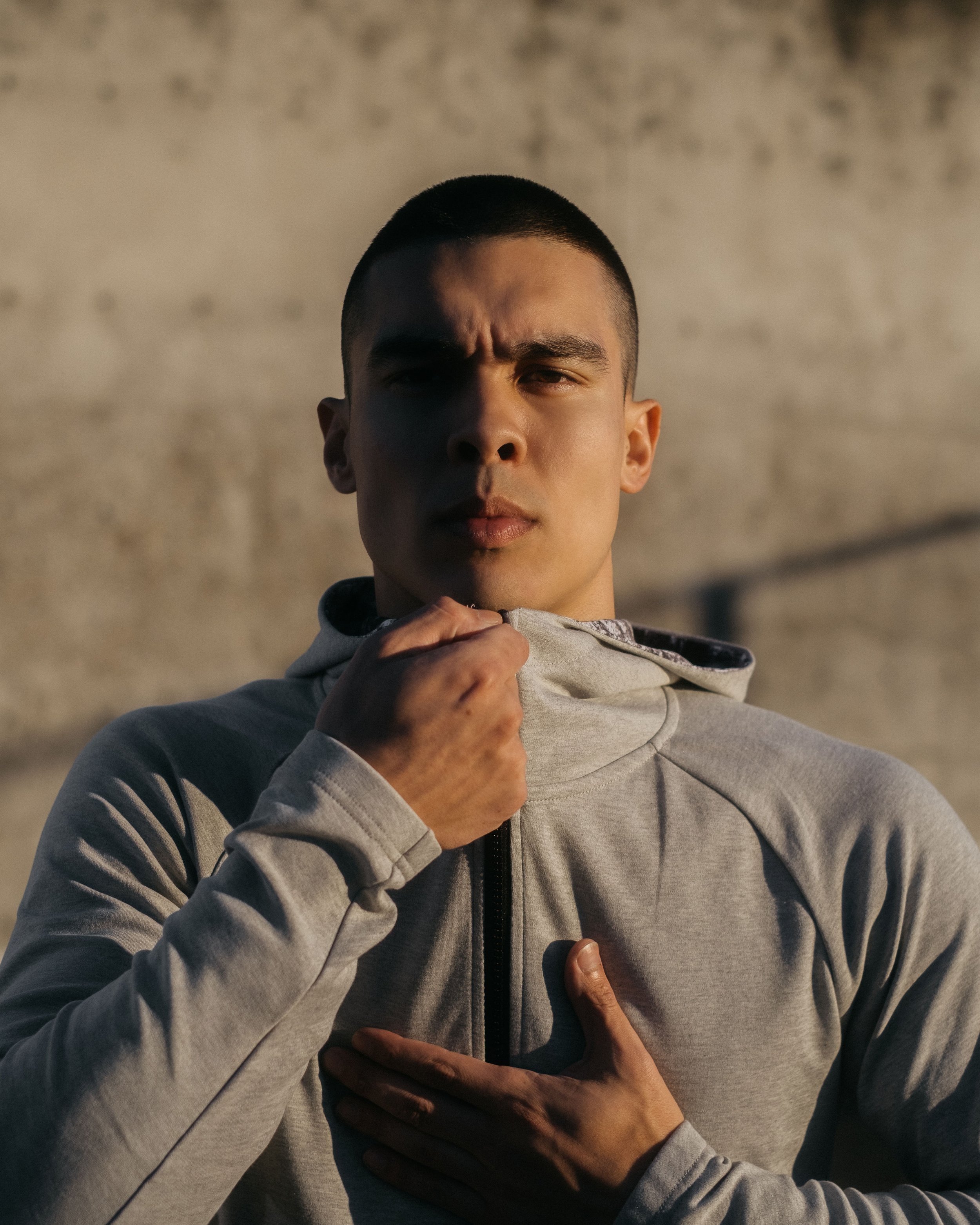 A young man in a gray athletic jacket touching his chest with one hand and holding the collar with the other, looking serious against a concrete background.