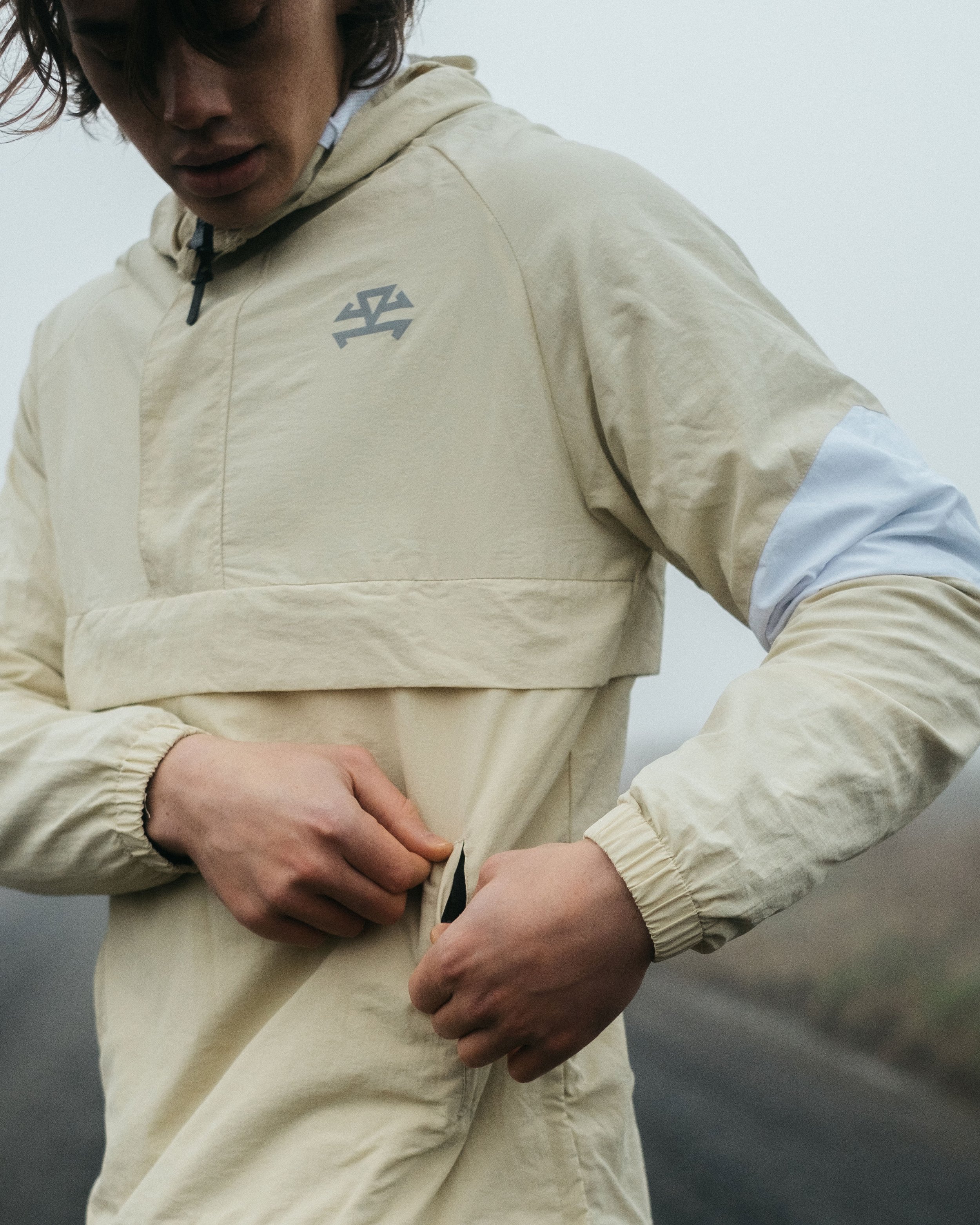 Person wearing a beige outdoor jacket with a logo on the chest, adjusting the waistband of their pants in a foggy outdoor setting.