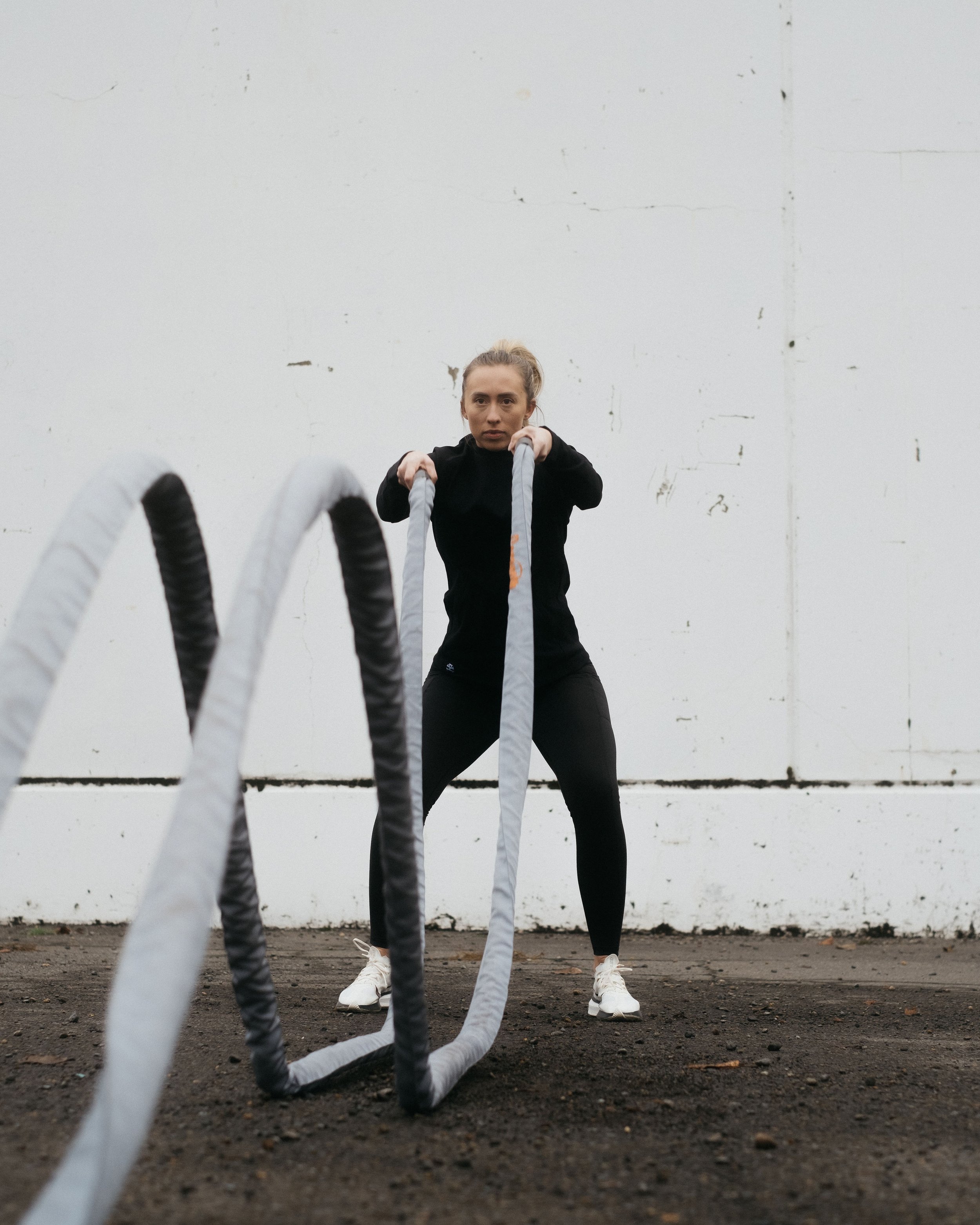 A woman in black workout clothes is holding battle ropes in an outdoor fitness area with a white wall behind her.