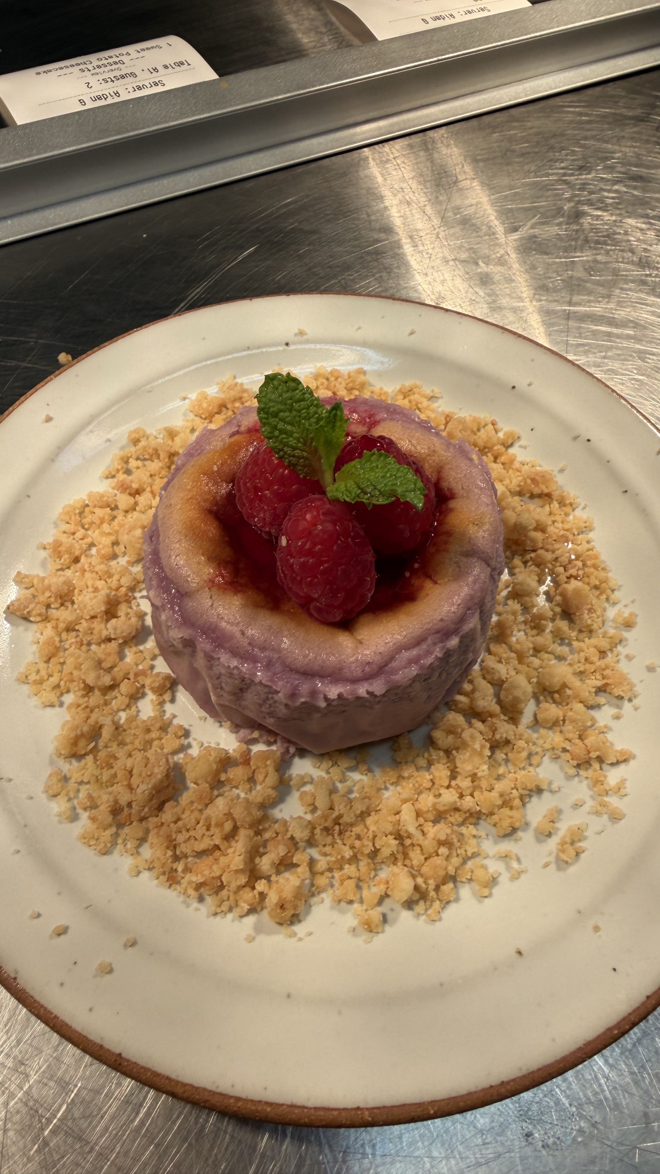A small, round dessert with a purple glaze, topped with raspberries and mint leaves, served on a white plate with crushed crumbles around it.