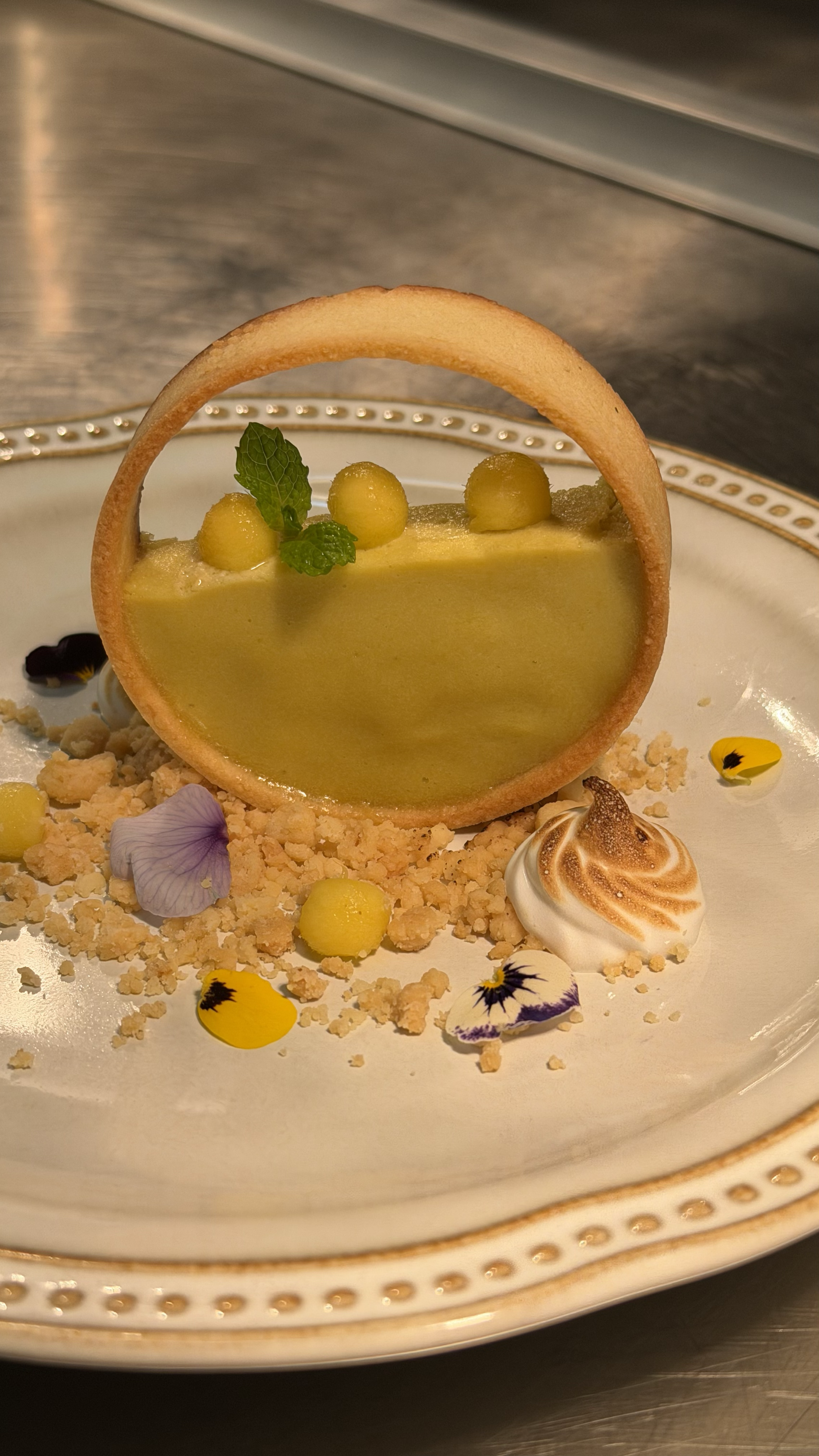 A dessert featuring a lemon tart with a circular cookie crust, garnished with a mint leaf, edible flowers, a dollop of meringue, and yellow spherical decorations, all on a decorative white plate.