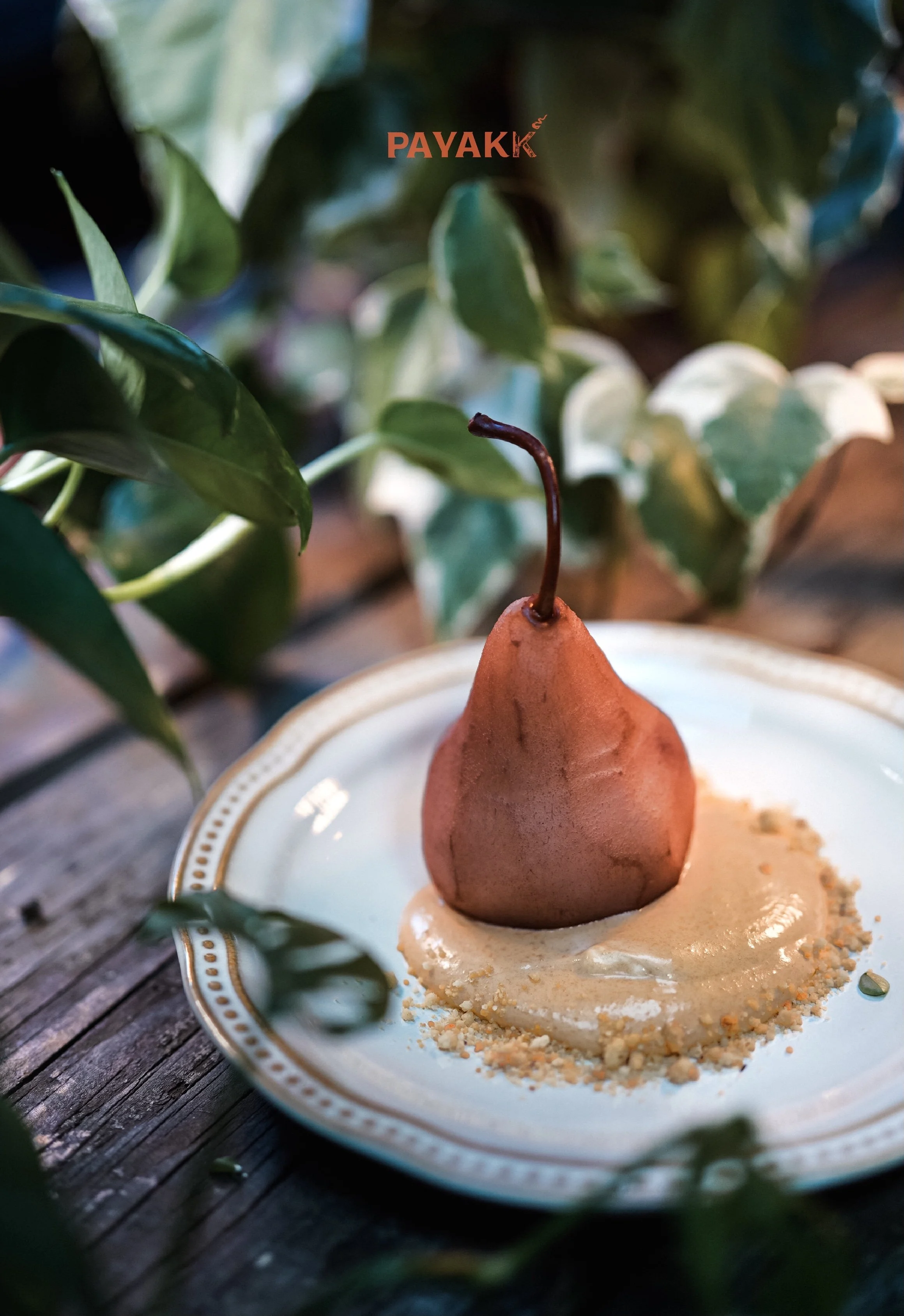 A pear-shaped dessert with a stem, placed on a decorative ceramic plate with a crumbly yellow substance around it. Green foliage in the background, and the word 'PAYAK' is written in red at the top of the image.