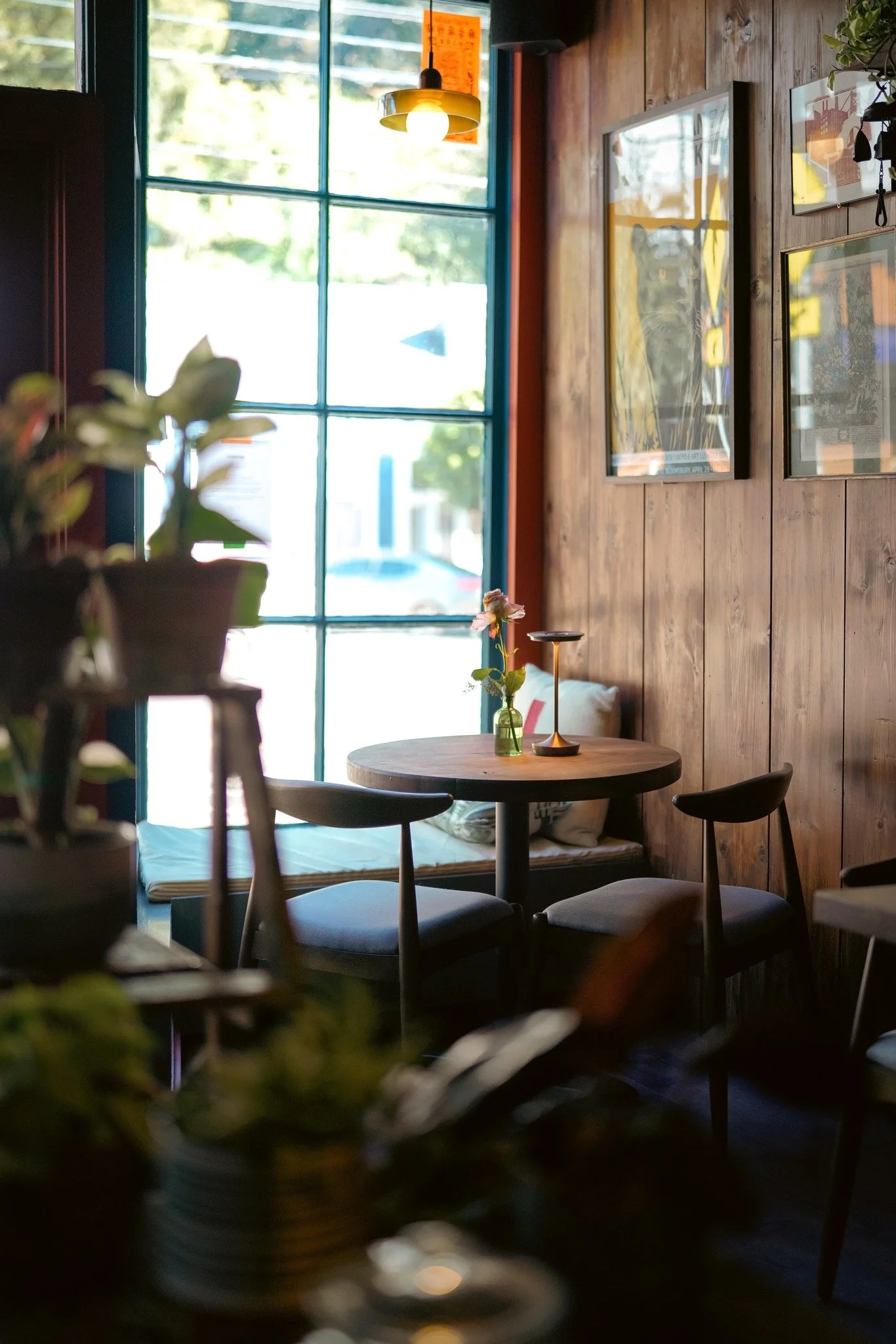 A cozy corner of a coffee shop with a round wooden table, two chairs, a window with green frames, and framed artwork on a wooden wall, illuminated by a hanging ceiling light.