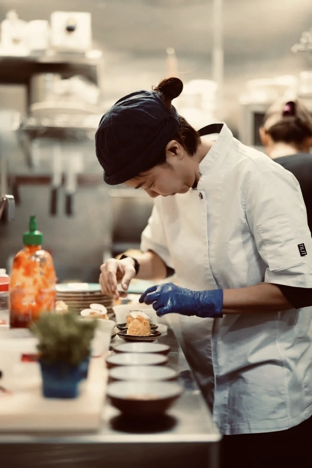 A female chef in a white uniform and black hat preparing food in a professional kitchen, focusing on plating dishes.