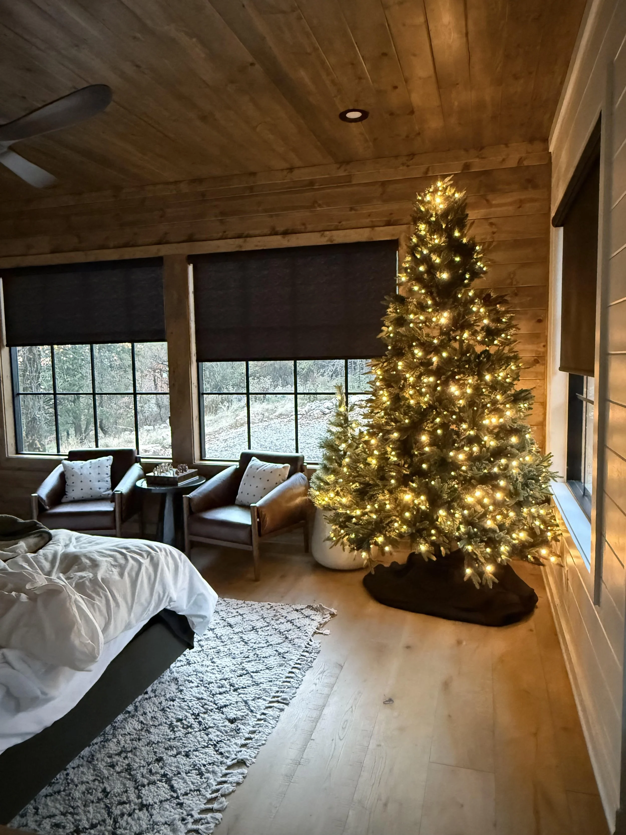 A cozy bedroom with a decorated Christmas tree illuminated with warm white lights next to a window, a bed with rumpled bedding, a patterned rug on wooden flooring, and two armchairs with pillows near the window.
