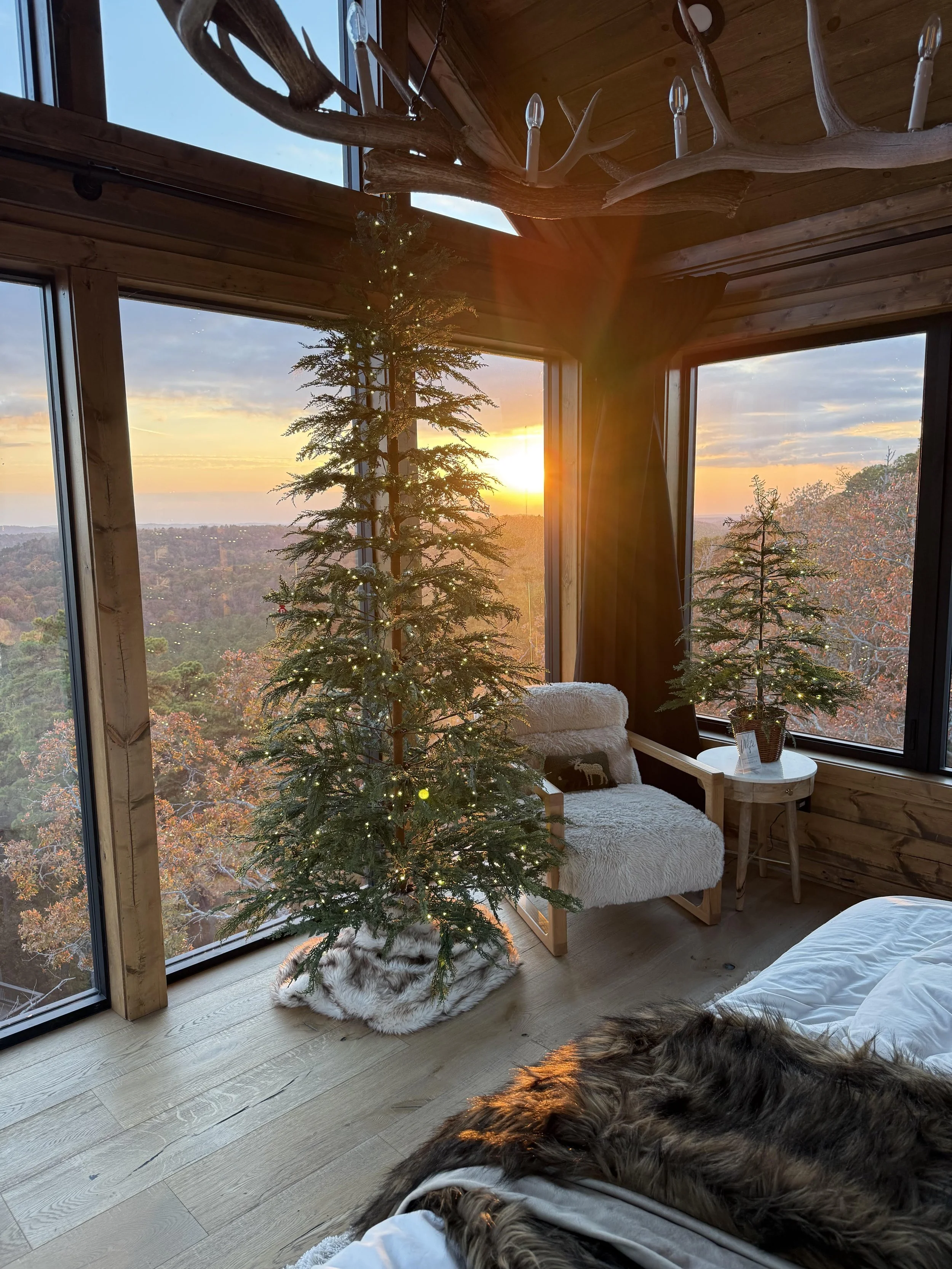 A cozy interior room with large windows showing a sunset over autumnal trees. A decorated Christmas tree with fairy lights stands beside the window, and a smaller tree with lights is on a table. An armchair with a fluffy throw blanket and a pillow is