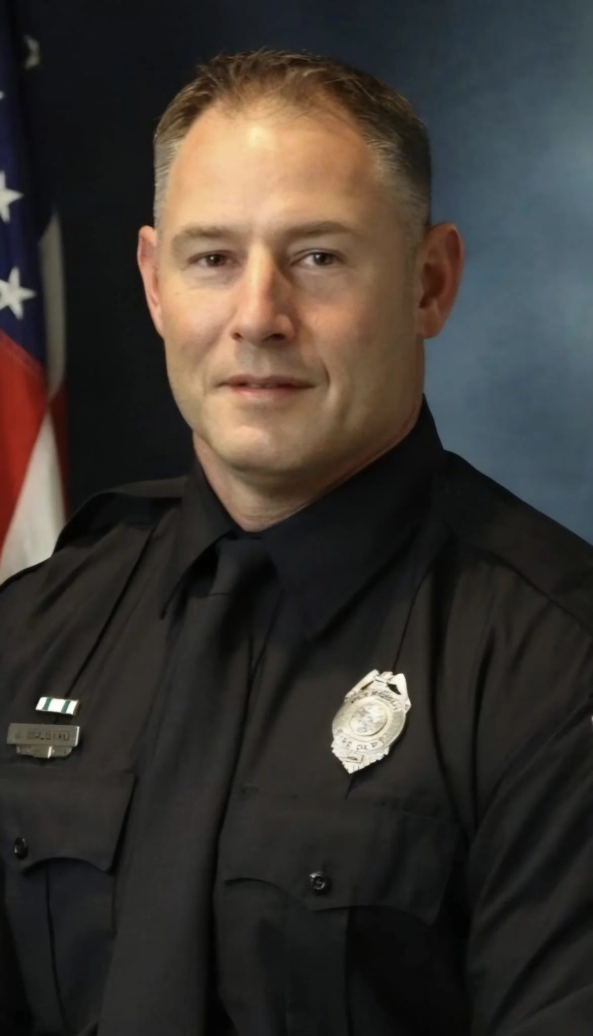 A male firefighter in a black uniform sitting with an American flag in the background. He has a badge and name tag on his uniform, and his arm patch indicates he is part of the Plymouth Fire-Rescue in Minnesota.