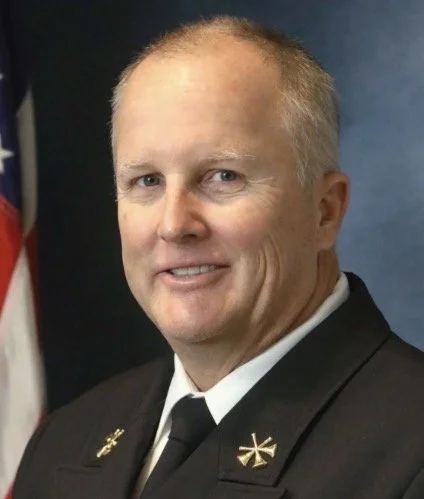 A male firefighter in uniform standing in front of an American flag.