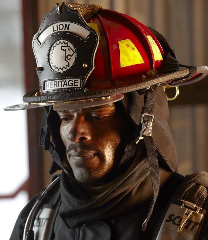 Firefighter wearing a helmet with a badge labeled "LION HERITAGE", with a firefighter uniform, standing indoors.