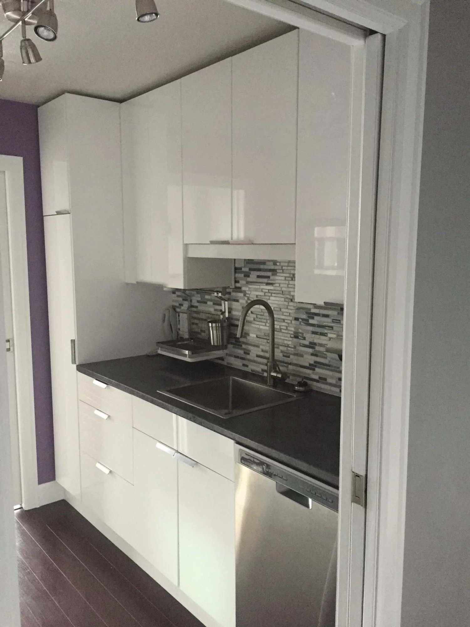 Small kitchen with white cabinets, dark countertop, mosaic tile backsplash, stainless steel sink, and dishwasher.