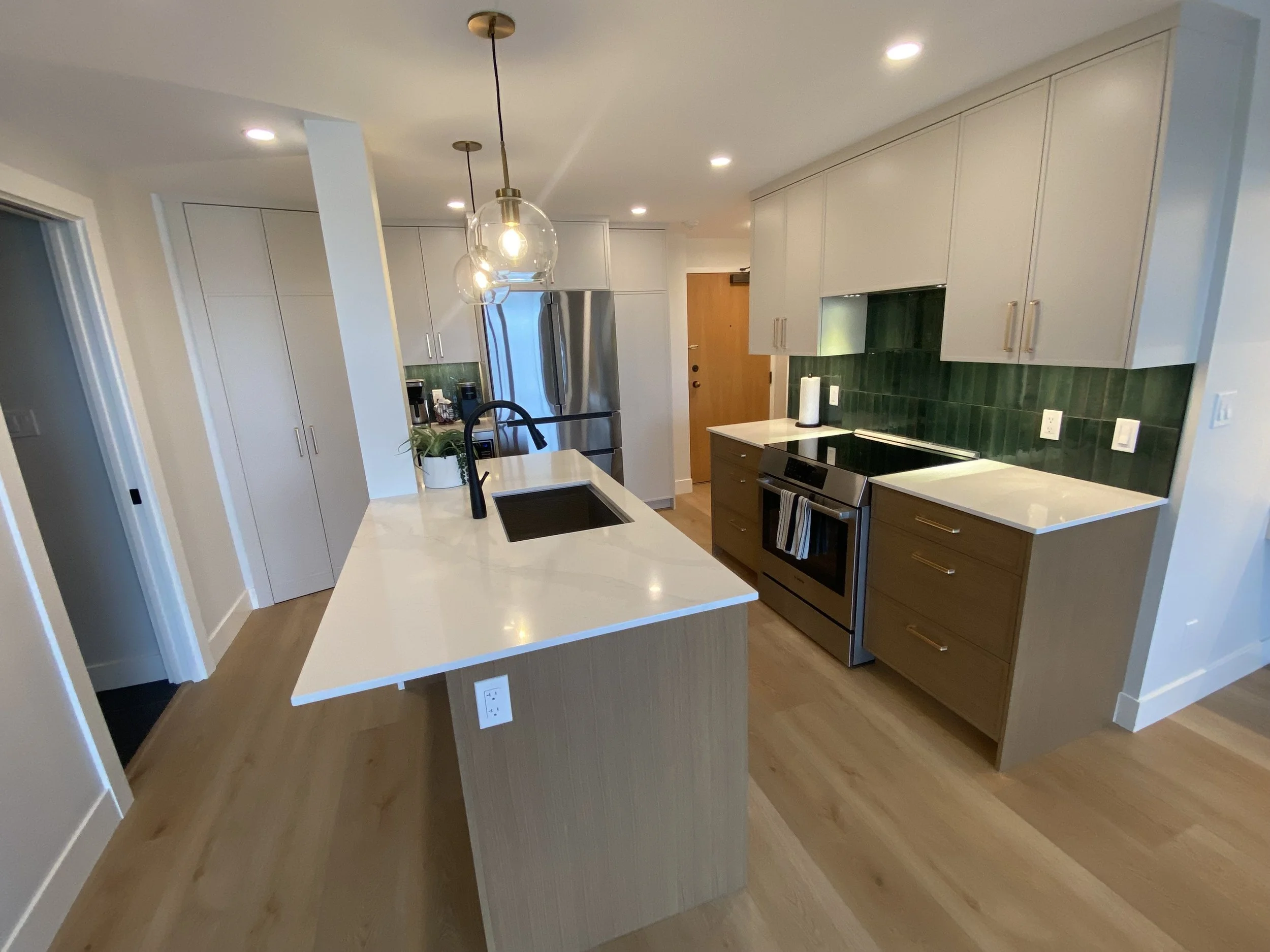 Modern kitchen with white cabinets, an island with a black sink and a pendant light, stainless steel refrigerator, oven, green tiled backsplash, and light wood flooring.