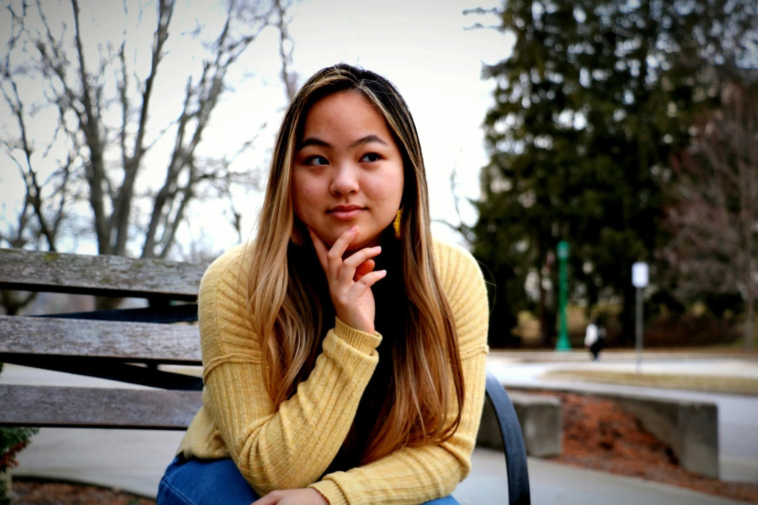 Asian female with blonde streaked brown hair and yellow sweater sitting on bench contemplating polyamory therapy near Concord, NC