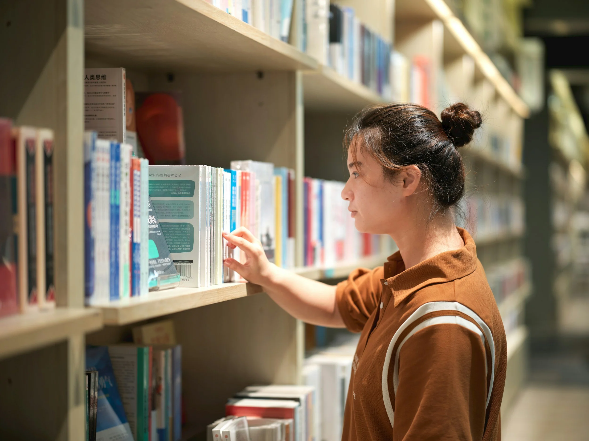 nonbinary individual with brown hair in bun and colored orange shirt looking through books on shelf