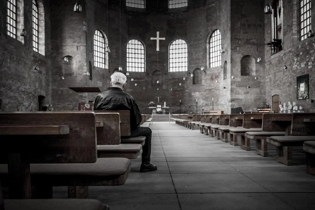 elder male with white hair and dark clothing sitting on pew in empty church considering religious trauma therapy in Concord, NC