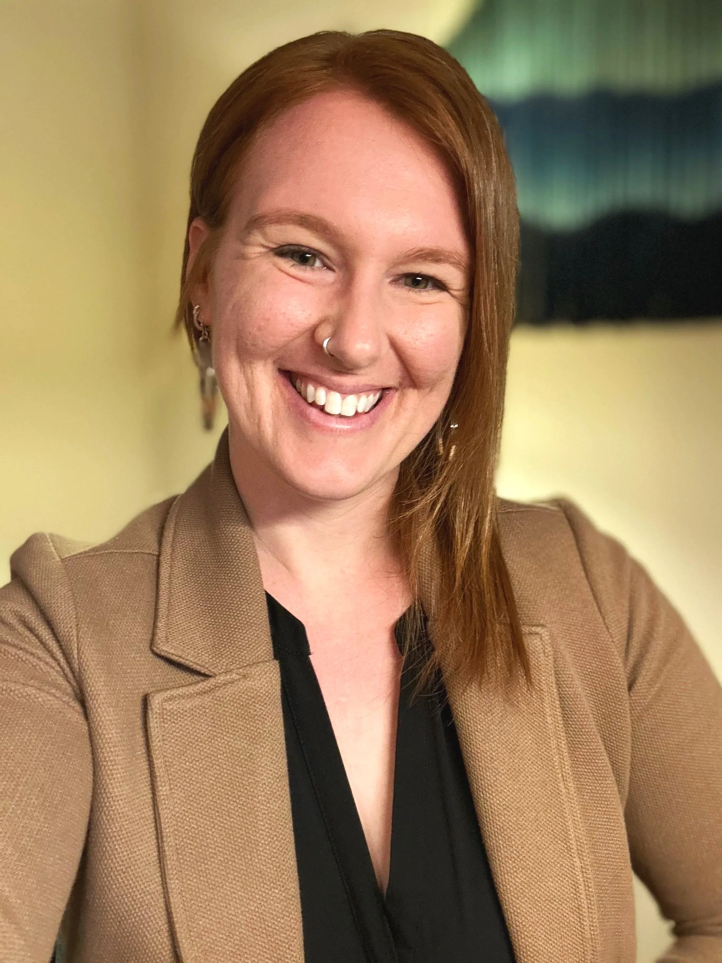 A woman trauma therapist with red hair smiling, wearing a brown blazer and earrings, indoors with yellow walls