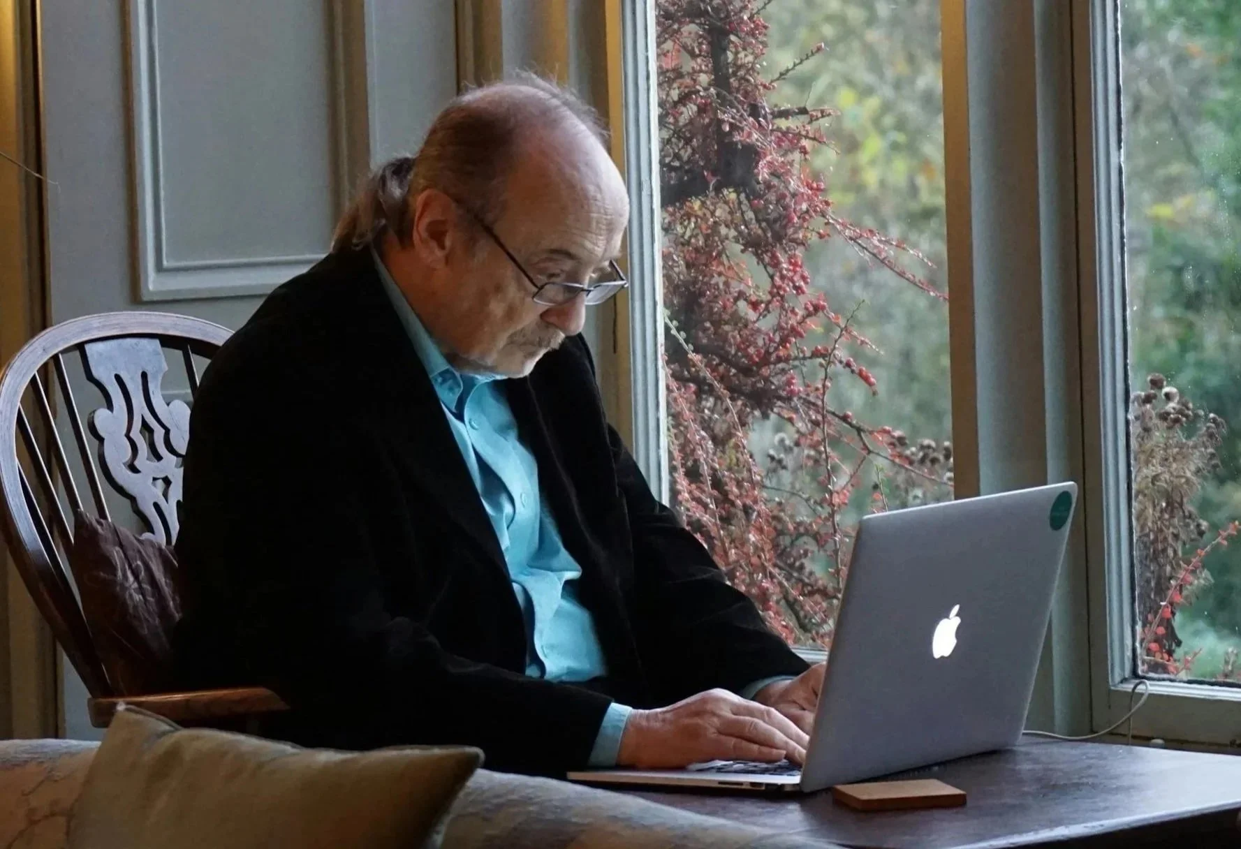 elder nonbinary client with glasses and ponytail sitting at desk by window on trauma therapy video appointment in Concord, NC