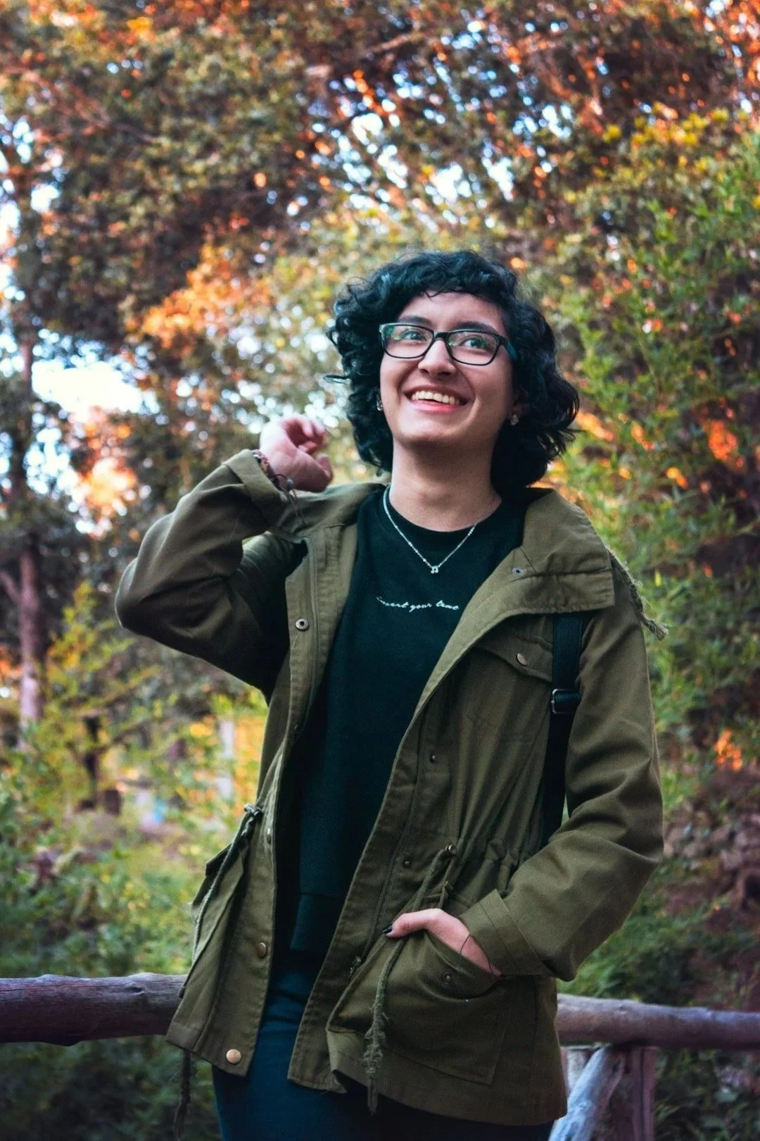 young adult with short dark curly hair and glasses wearing black shirt with green jacket smiling outside after religious trauma therapy in Concord, NC