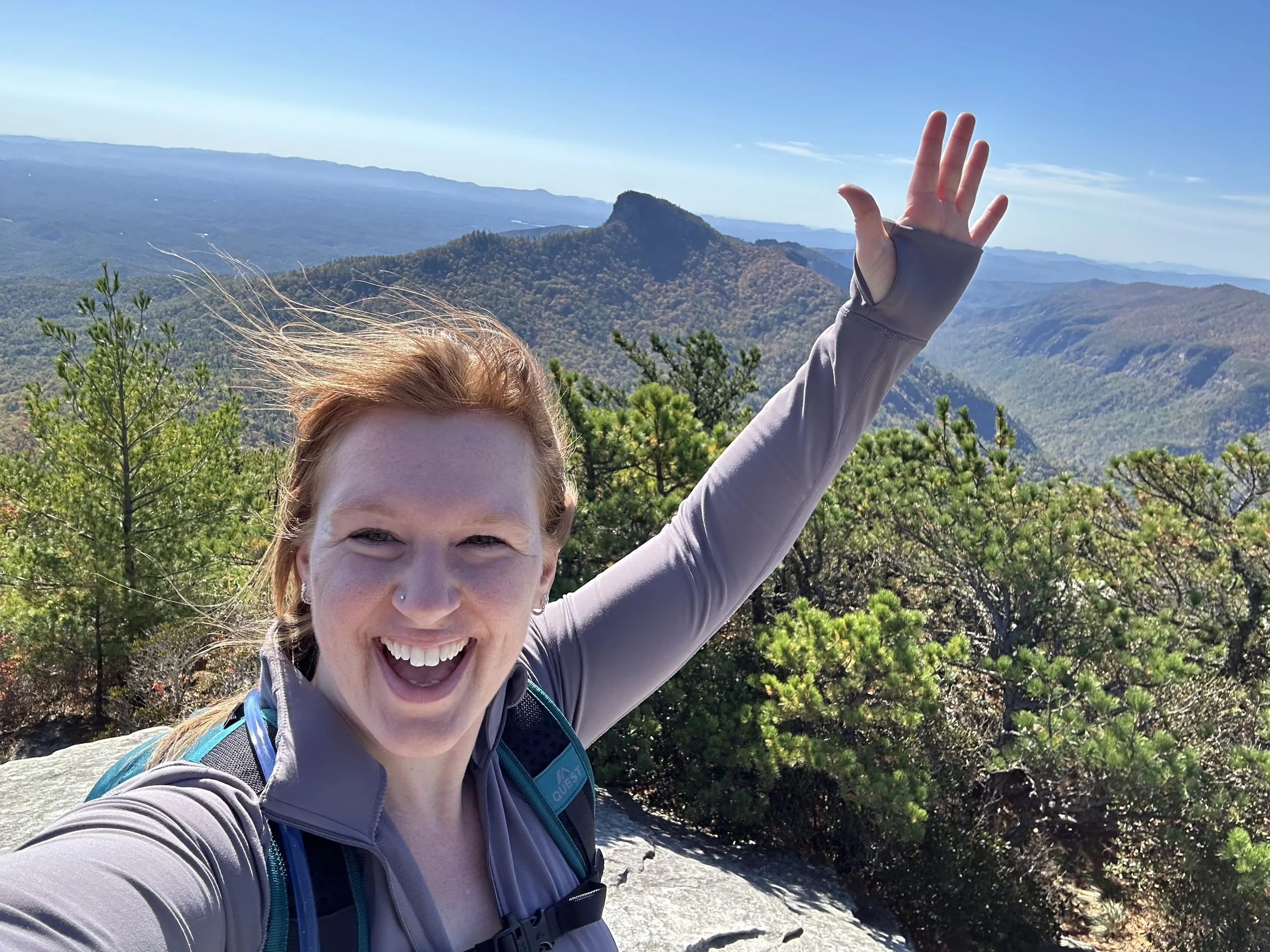 Trauma therapist standing on top of mountain smiling with arm raised and view of Blue Ridge in background near Concord, NC