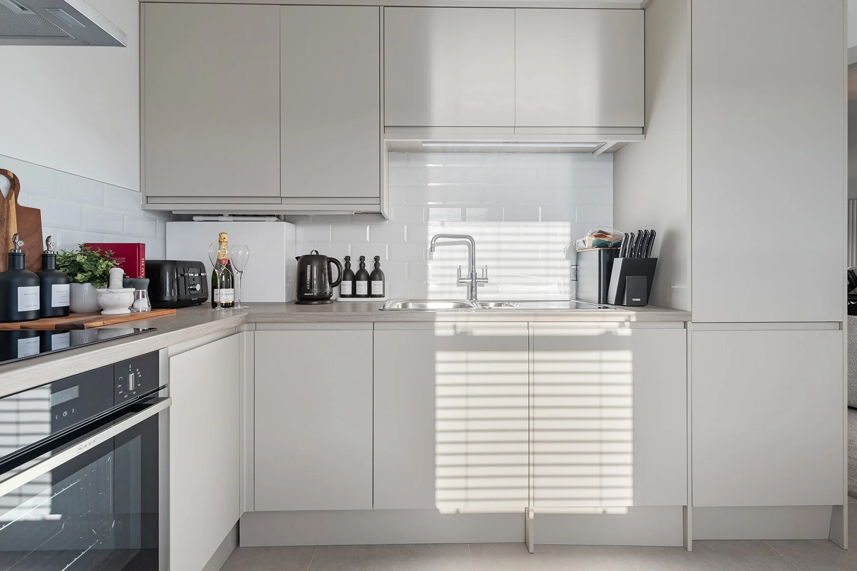 Modern living room with a white sofa, black and white pillows, a round wooden coffee table with decorative items, a contemporary beige armchair with a pillow and throw blanket, a tall potted plant, a wooden chair, and a floor lamp, all on a light-colored rug with dark wood flooring.