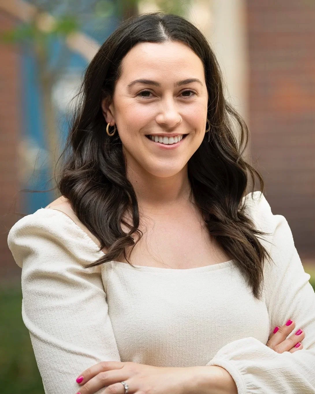 Headshot of Carolyn Levine, a young woman with long dark wavy hair, wearing a cream-colored top, smiling with folded arms, standing outdoors with blurred background of trees and building.