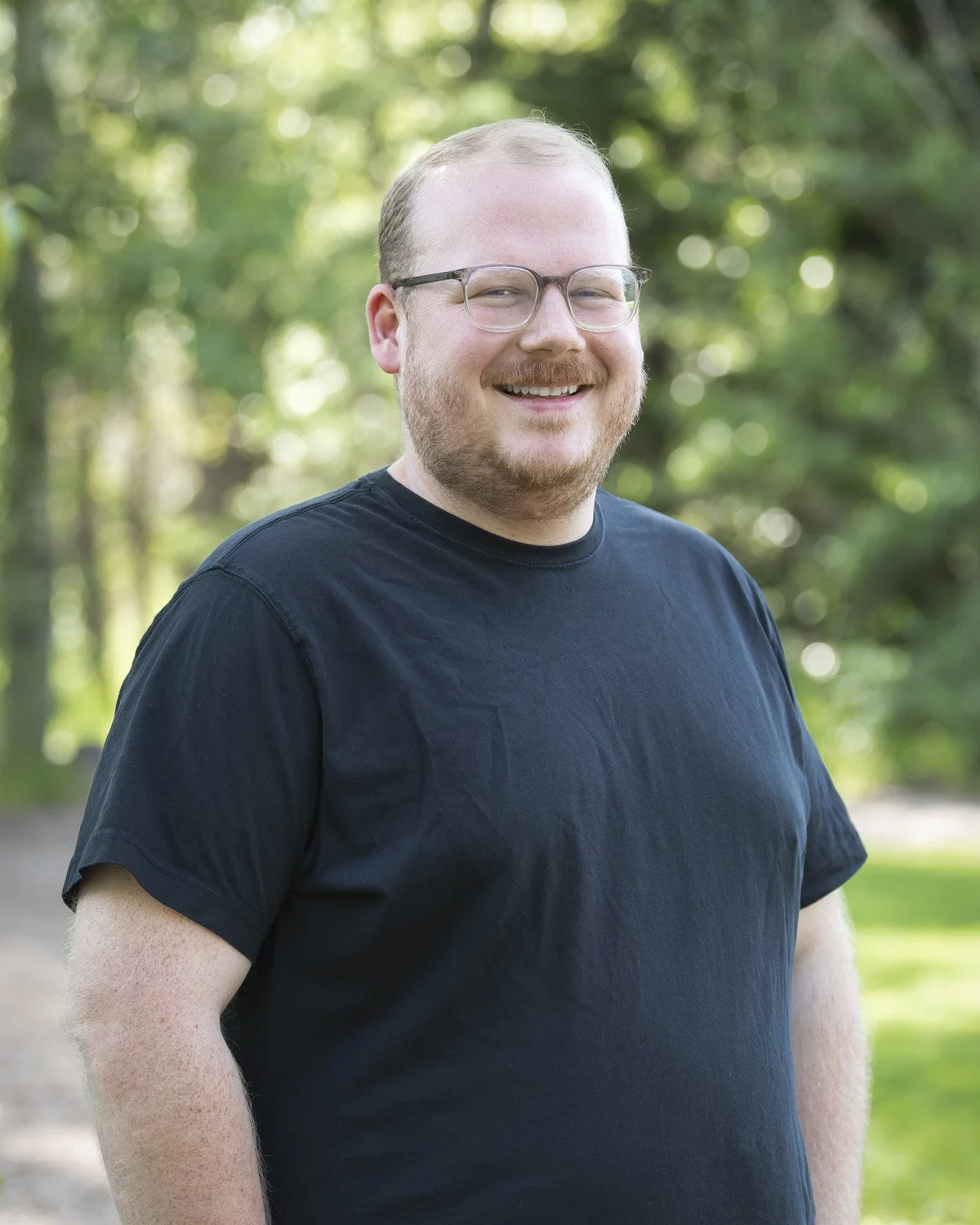 Headshot of Sam Daniel, a smiling man wearing glasses and a black t-shirt outdoors with trees and greenery in the background.