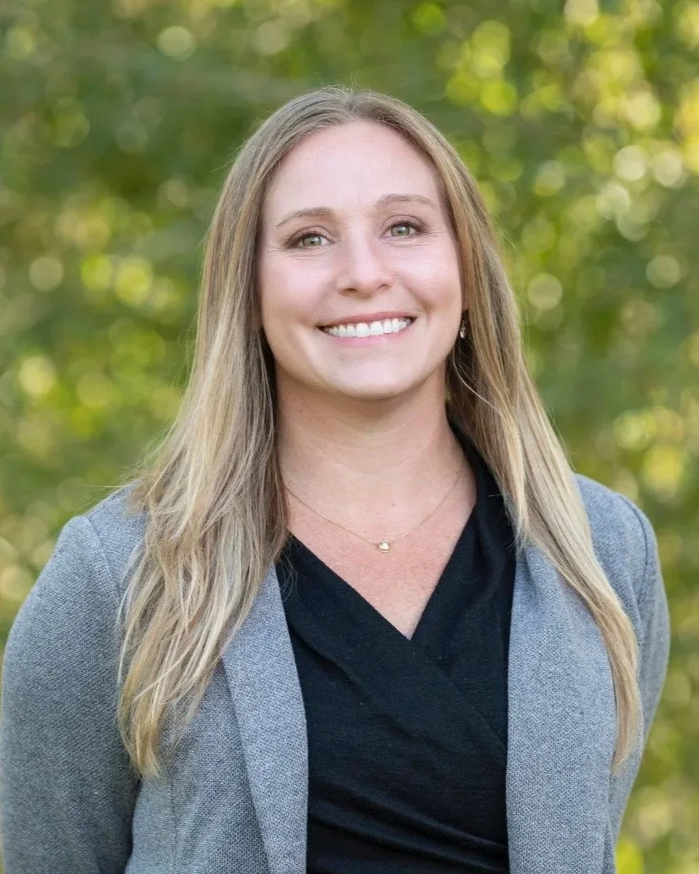 Headshot of Alison Cotey, a smiling woman with long blonde hair, wearing a gray blazer and black top, standing outdoors with a blurred green background.