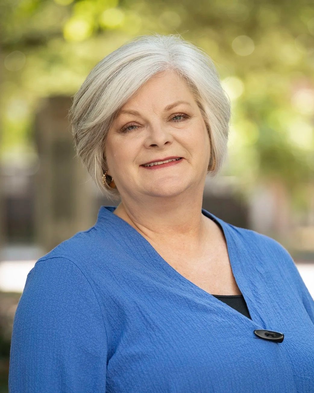 Portrait of Grace Tice, an elderly woman with short gray hair, wearing a blue top, smiling softly outdoors with a blurred green background.