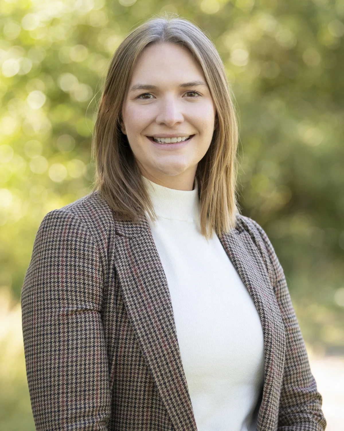 Headshot of Emily Pixberg, a young woman with shoulder-length light brown hair, smiling outdoors with blurred green trees in the background, wearing a white turtleneck and a brown checked blazer.