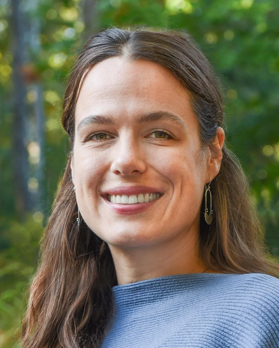 Headshot of Julia Prince, a close-up of a smiling woman with long brown hair, wearing earrings and a blue top, outdoors in a green forested area.