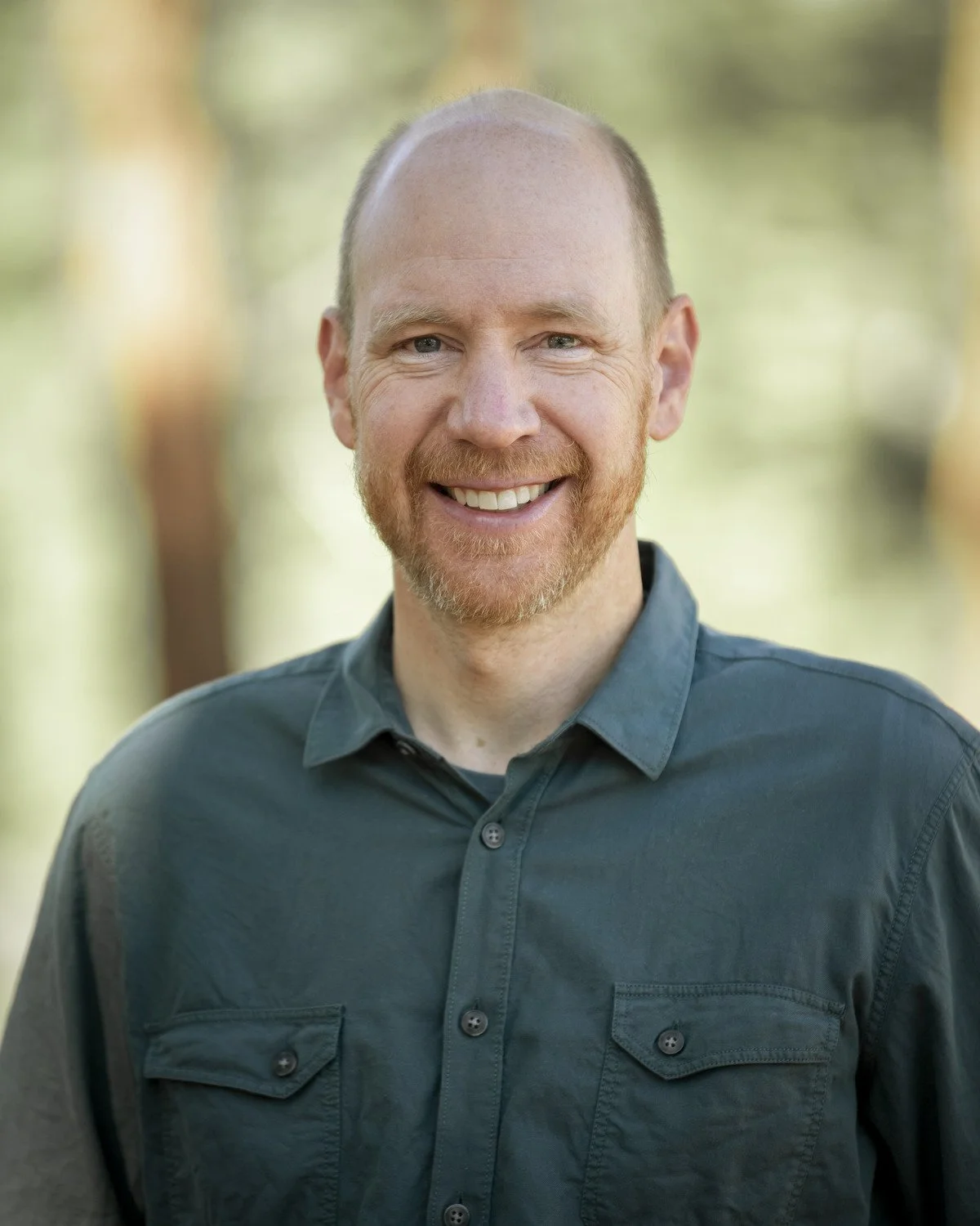 Headshot of Ben Fish, a smiling man with a beard and short hair wearing a dark teal button-up shirt outdoors with blurred trees in the background.