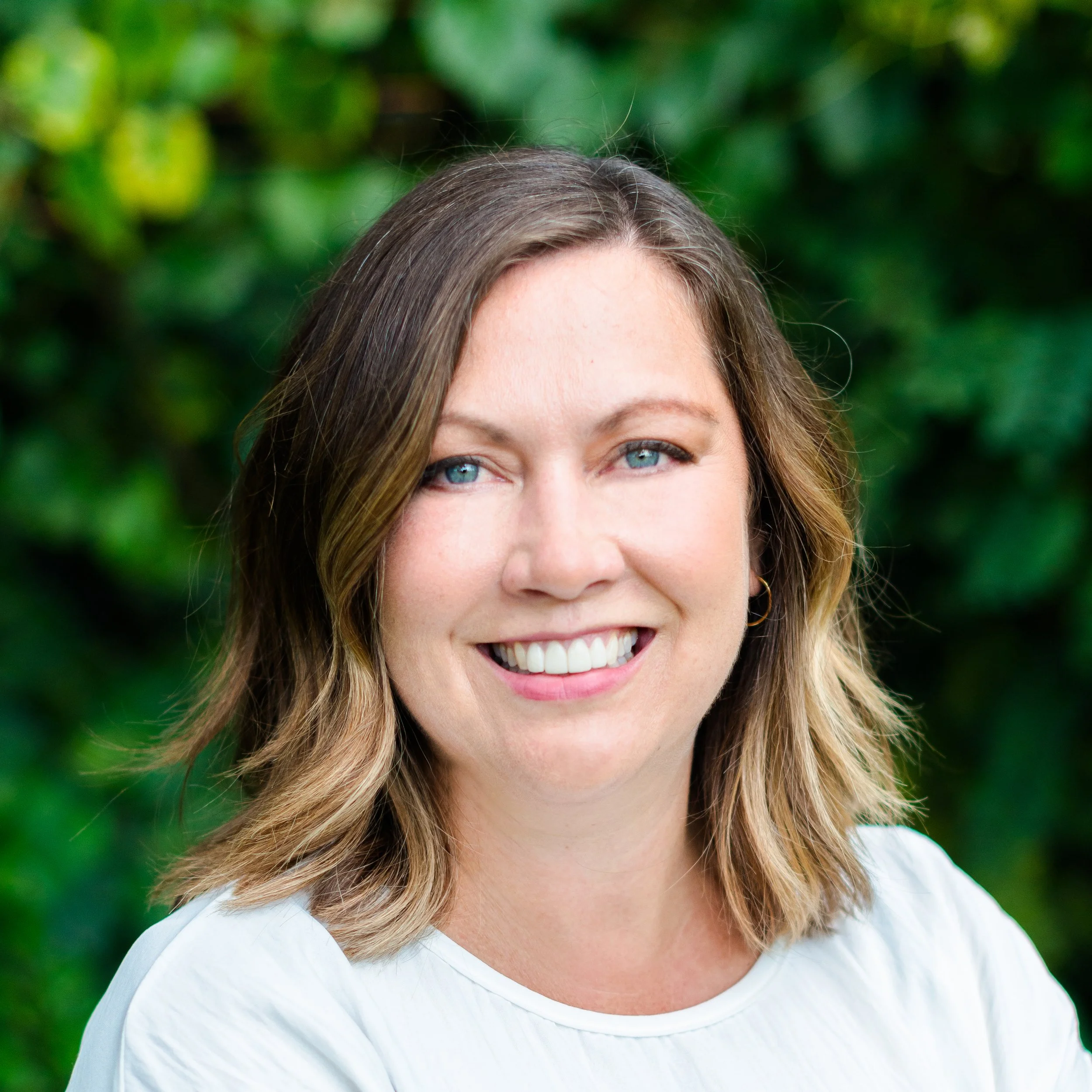 Headshot of Emily McCoy, a smiling woman with shoulder-length light brown hair and blue eyes, wearing a white top, standing outdoors against a blurred green foliage background.