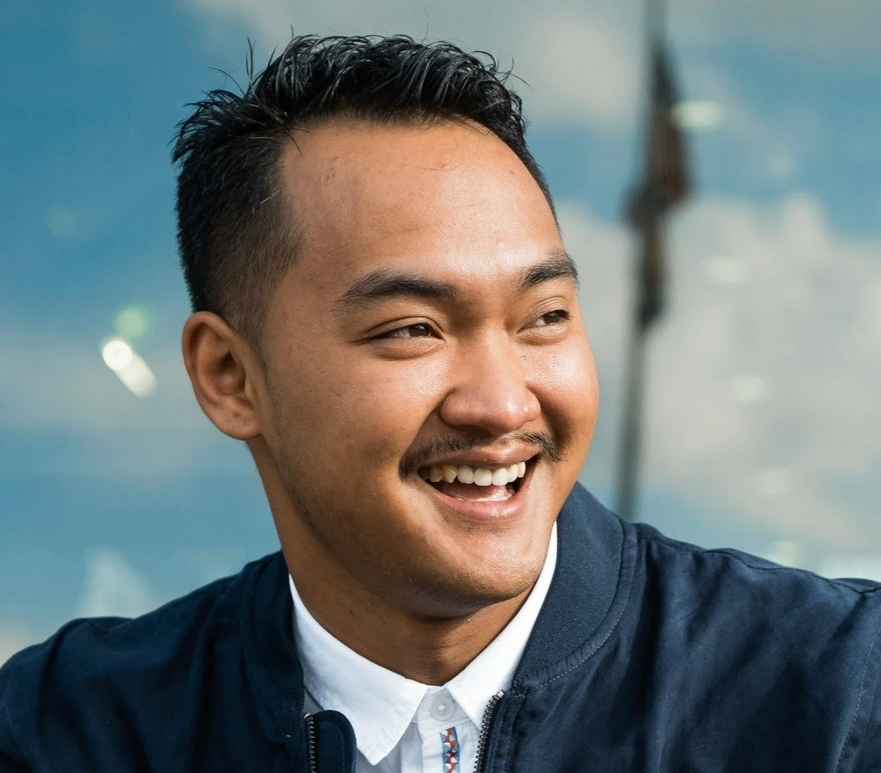 Headshot of Mo Bachtiar, a smiling young man with short black hair and a mustache, wearing a navy jacket over a white shirt, looking to the side with a cheerful expression.