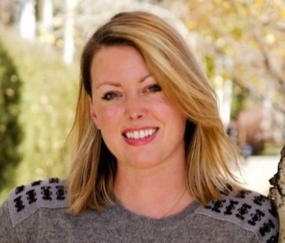 Headshot of Rielly Thimmons, a woman with shoulder-length blonde hair wearing a gray shirt, smiling outdoors with trees and autumn leaves in the background.
