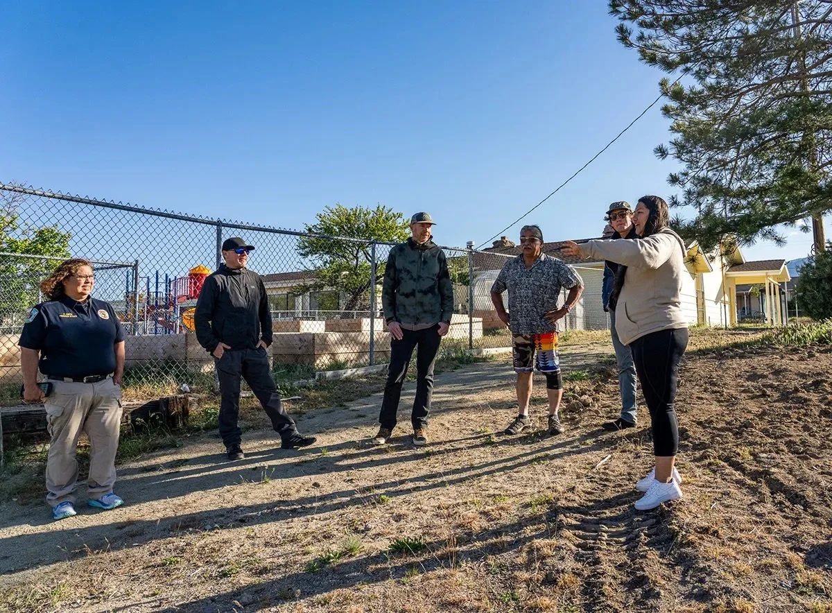 Group of six people standing outdoors, listening to a woman gesturing and talking, next to a fence with trees and houses in the background.