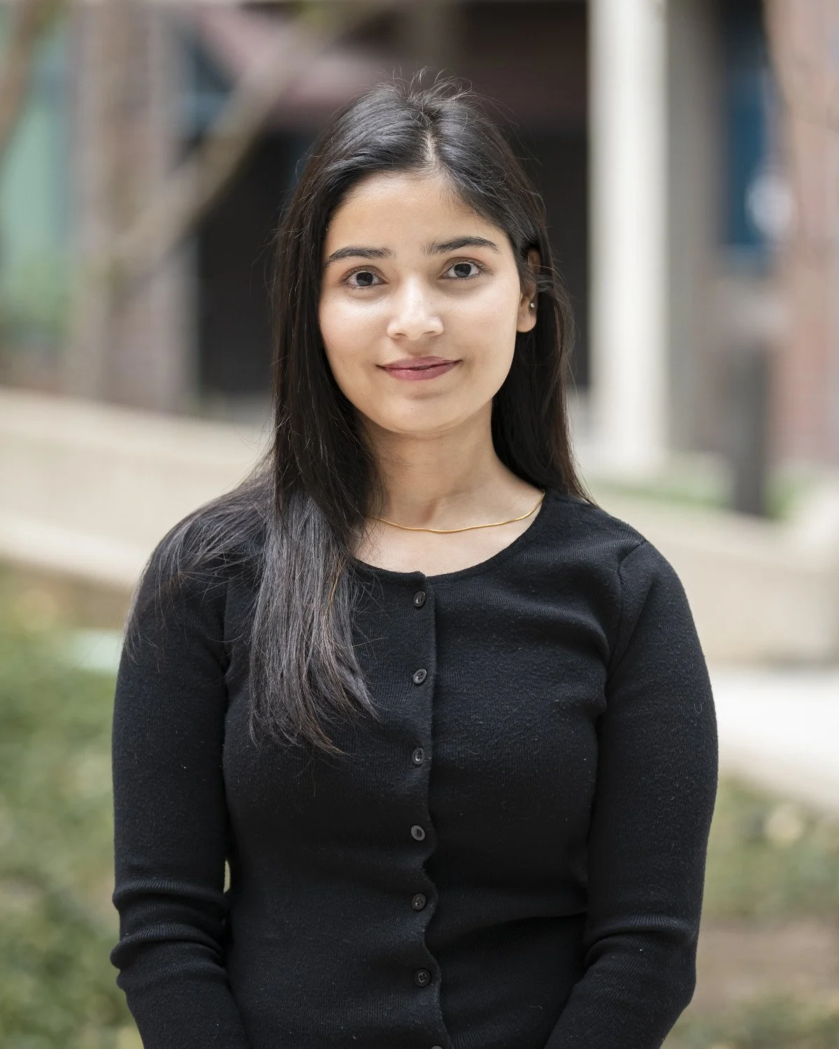Headshot of Samata Gyawali, a young woman with long dark hair and fair skin, wearing a black button-up cardigan and a thin gold necklace, standing outdoors in front of a blurred background.