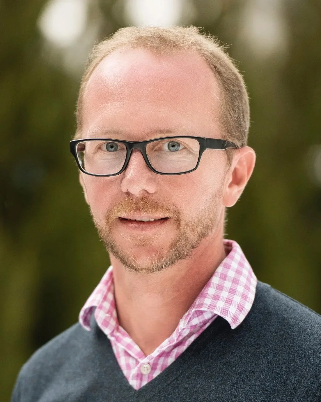 Headshot of Chris Geddes, a man with light skin, glasses, light brown hair, and a beard, wearing a pink checkered shirt and a dark sweater, outdoors with a blurred natural background.
