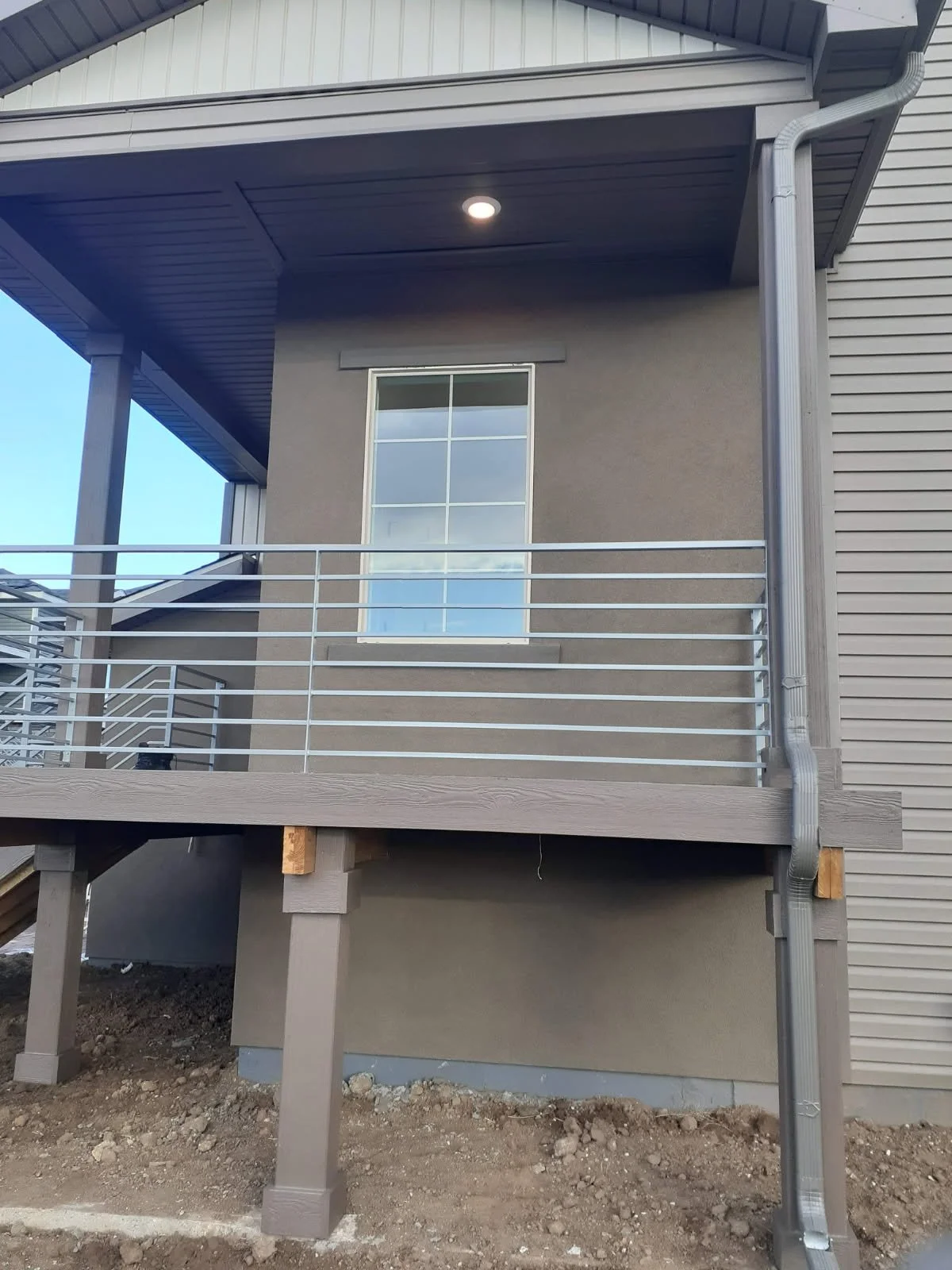 The photo shows the exterior of a house with a second-story porch, a window with a grid pattern, metal railing, and visible construction supports. The ground is bare dirt, and the house has siding and gutter systems.